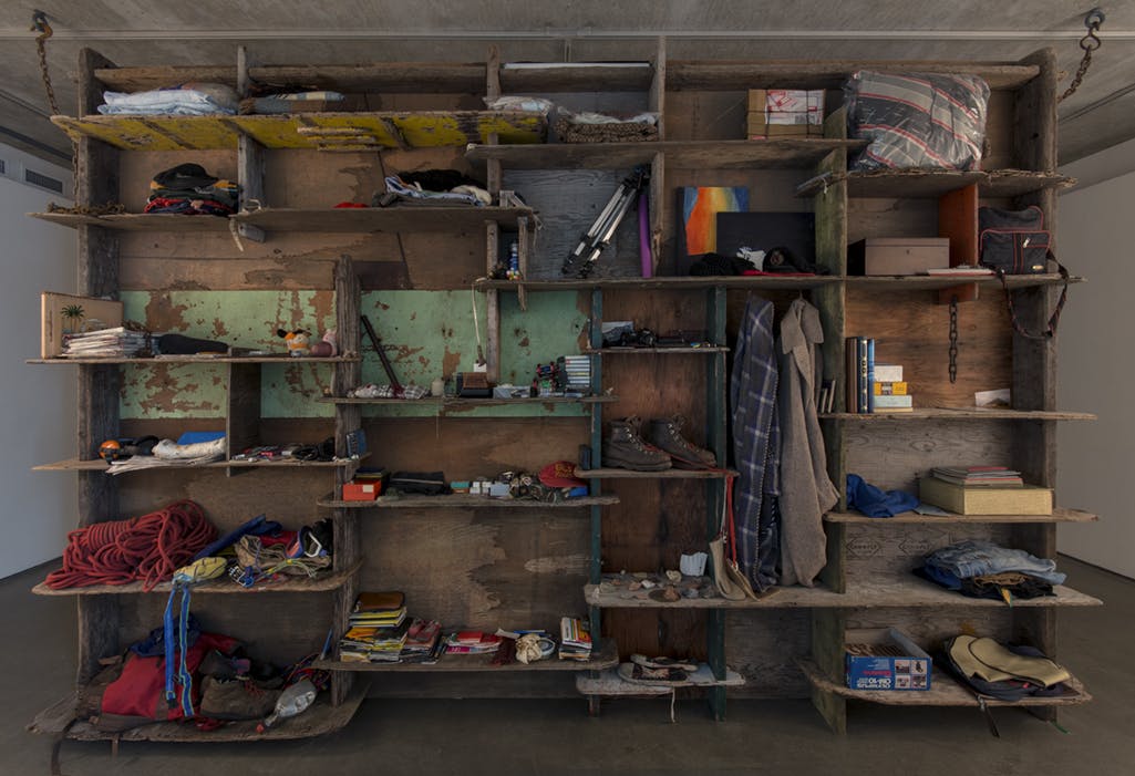 Large, rough wooden shelves are installed in a gallery space. Many objects such as a box,  clothes, a stack of rope, shoes, a shoulder bag, and notebooks are piled up on these shelves. 