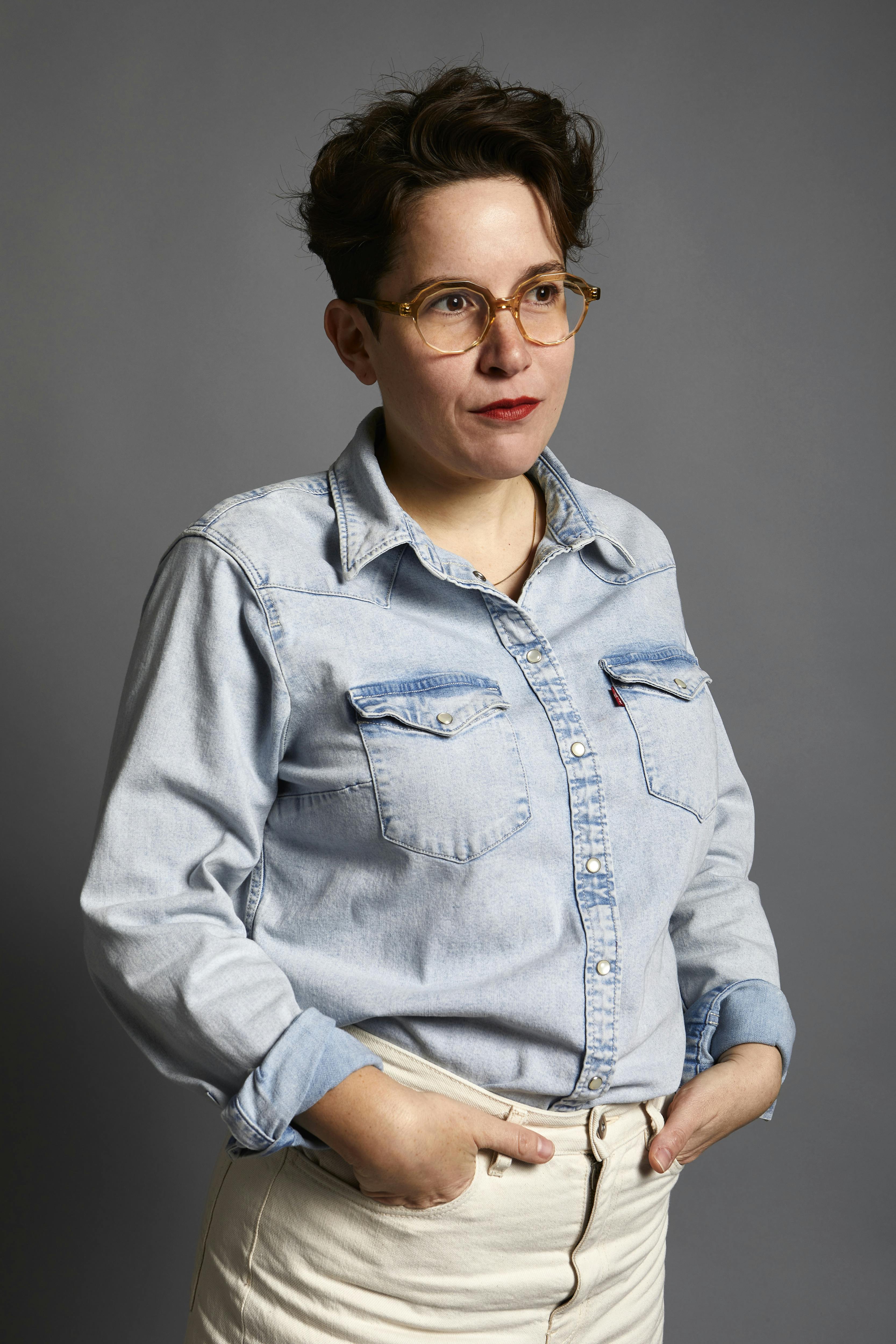 A portrait of Allison Grimaldi Donahue. She is wearing glasses, a light-blue denim button up and white pants.