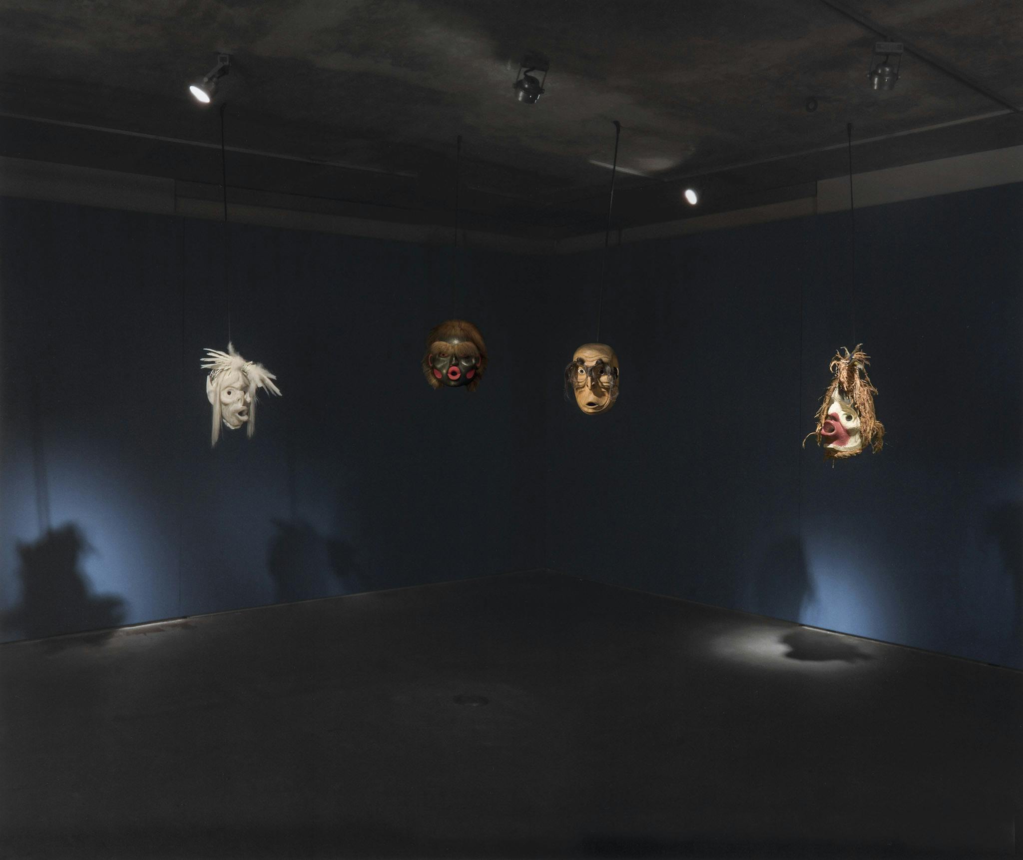 Installation image of four carved wood masks made by Beau Dick hanging in a gallery space. Each mask shows different faces with different facial expressions. The space is dimly lit and the walls are painted dark blue.