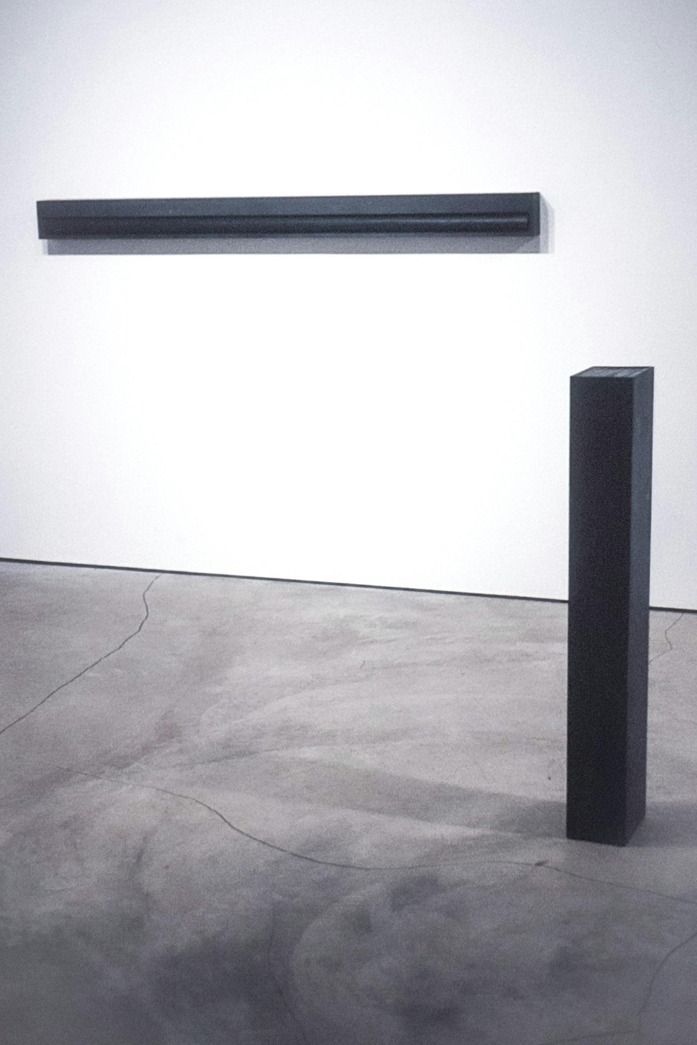 2 artworks in a gallery. One is a black rectangular prism with a black cylinder embedded in it, mounted horizontally on the wall. The other is a black rectangular prism placed vertically on the floor. 