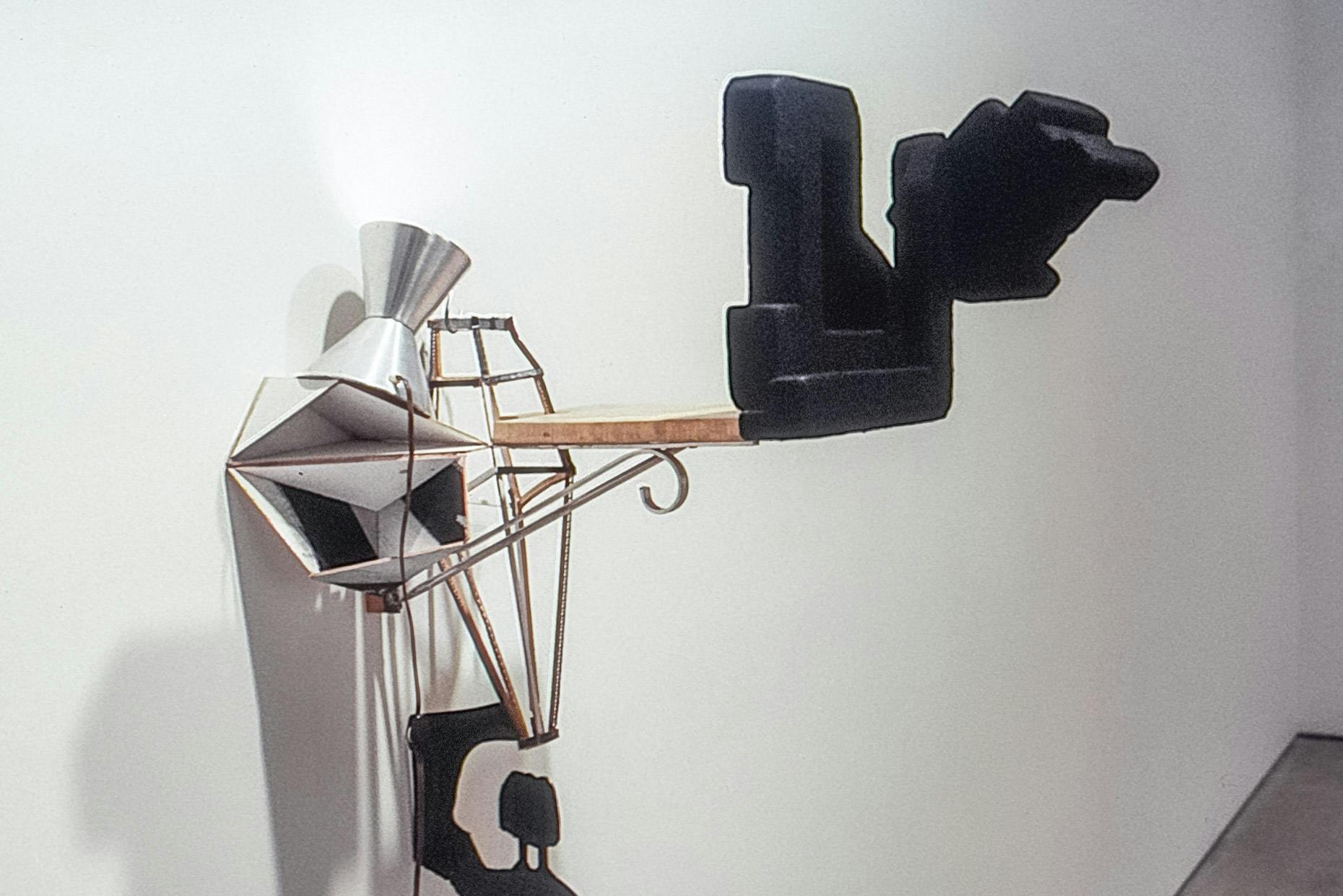 A closeup of a sculpture mounted on a white wall. The sculpture is made of many different items, including a double shot jigger, a small wood plank, a metal plant hook, and geometric black foam forms.