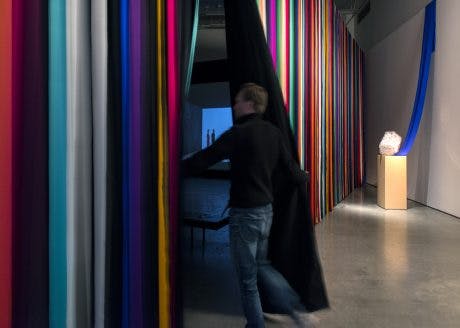 A striped multicoloured curtain hangs in a gallery. The curtain divides the space at an angle. A person opens the curtain to enter the other side, revealing a glimpse of a projection on the far wall. 