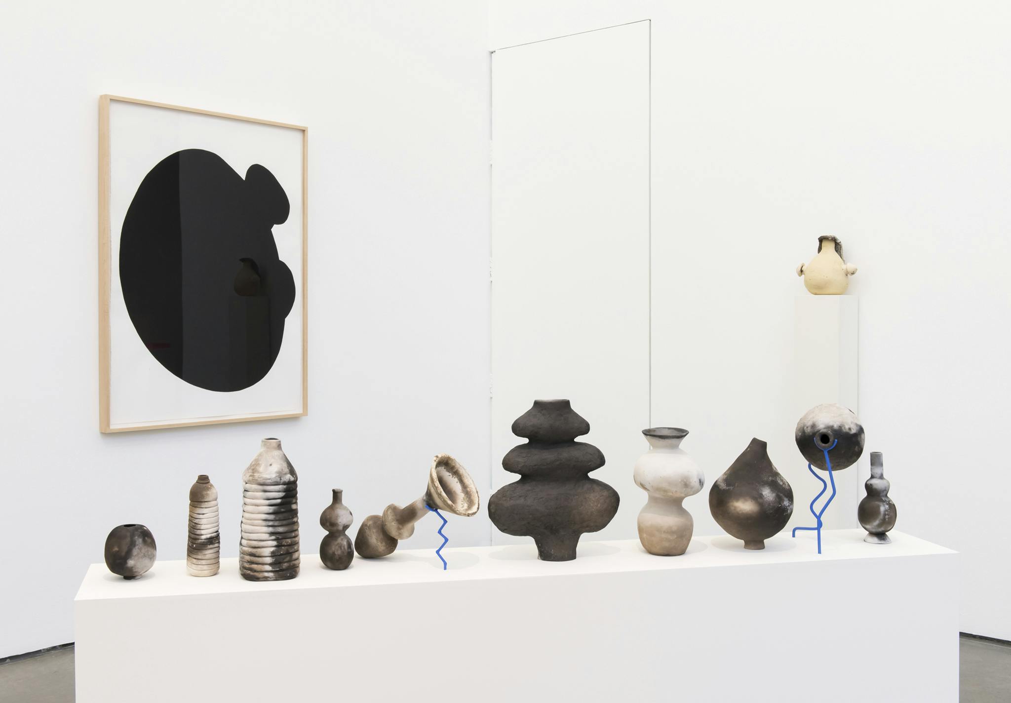 Ten ceramic sculptures of various shaped bottles sit on top of a long rectangular plinth. Behind the plinth, a large black and white painting hangs and another sculpture on a tall plinth to the right.