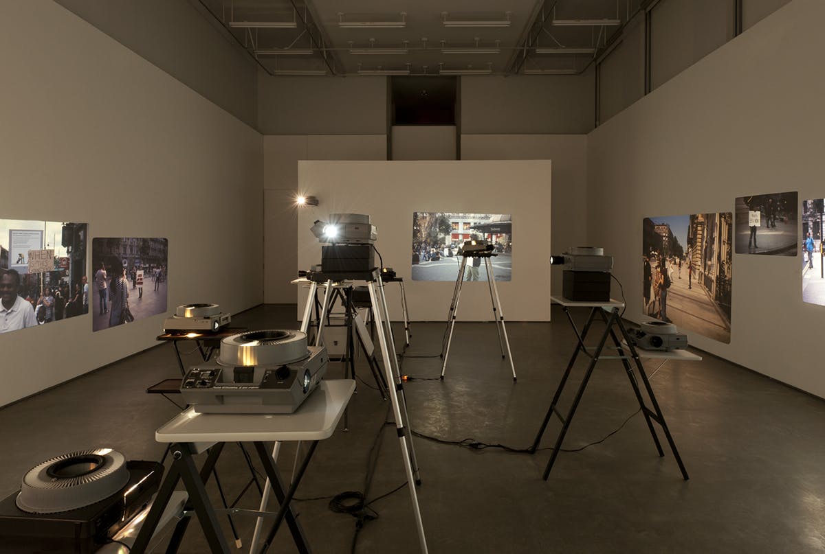 Eight slide projectors are visible in this image, each of which are on tables at various heights. They project photographs onto the three surrounding walls.