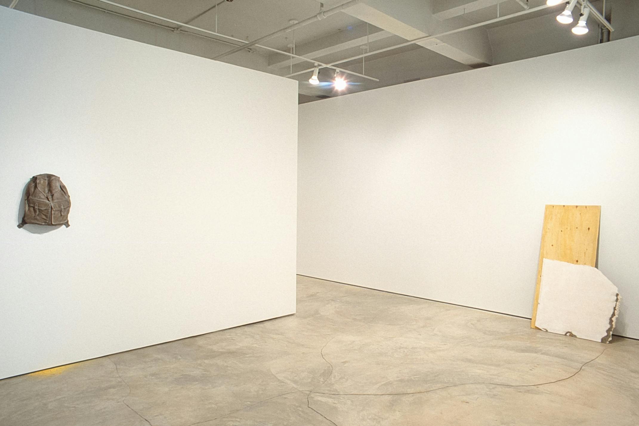 Two artworks are installed in the gallery space. One is a closed backpack with yellow paint scattered beneath where it is mounted. Thick paper and a piece of wood are leaning against the other wall.