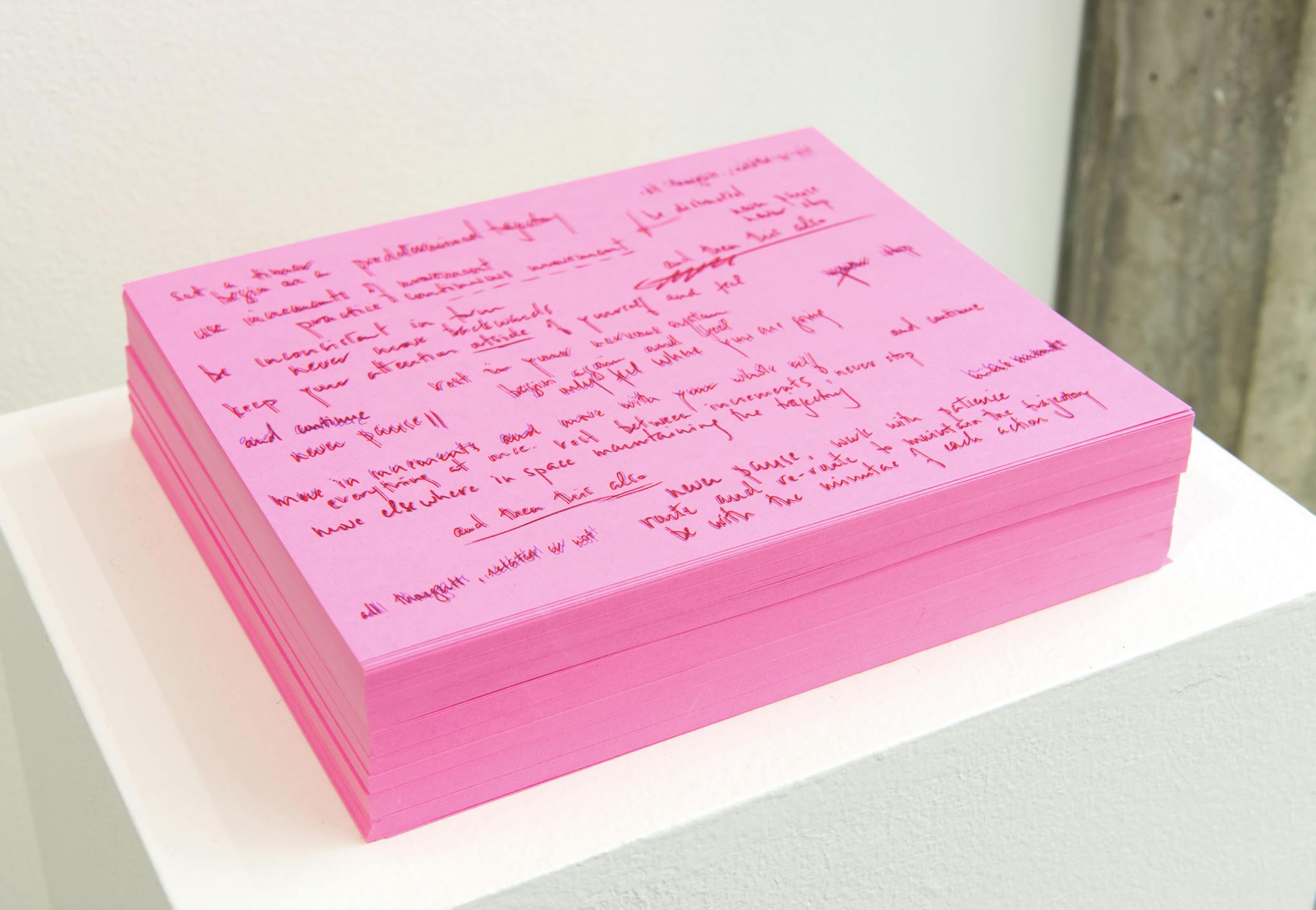 A stack of prints on pink paper sits on top of a white plinth. The prints are of handwritten notes scrawled across the page. 