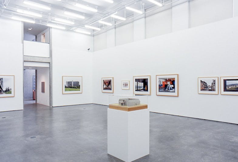 In a gallery, there are many colour photos of different sizes in wooden frames on the wall. in the centre, there is a white plinth with a glass case on top, holding a maquette of a low grey building.