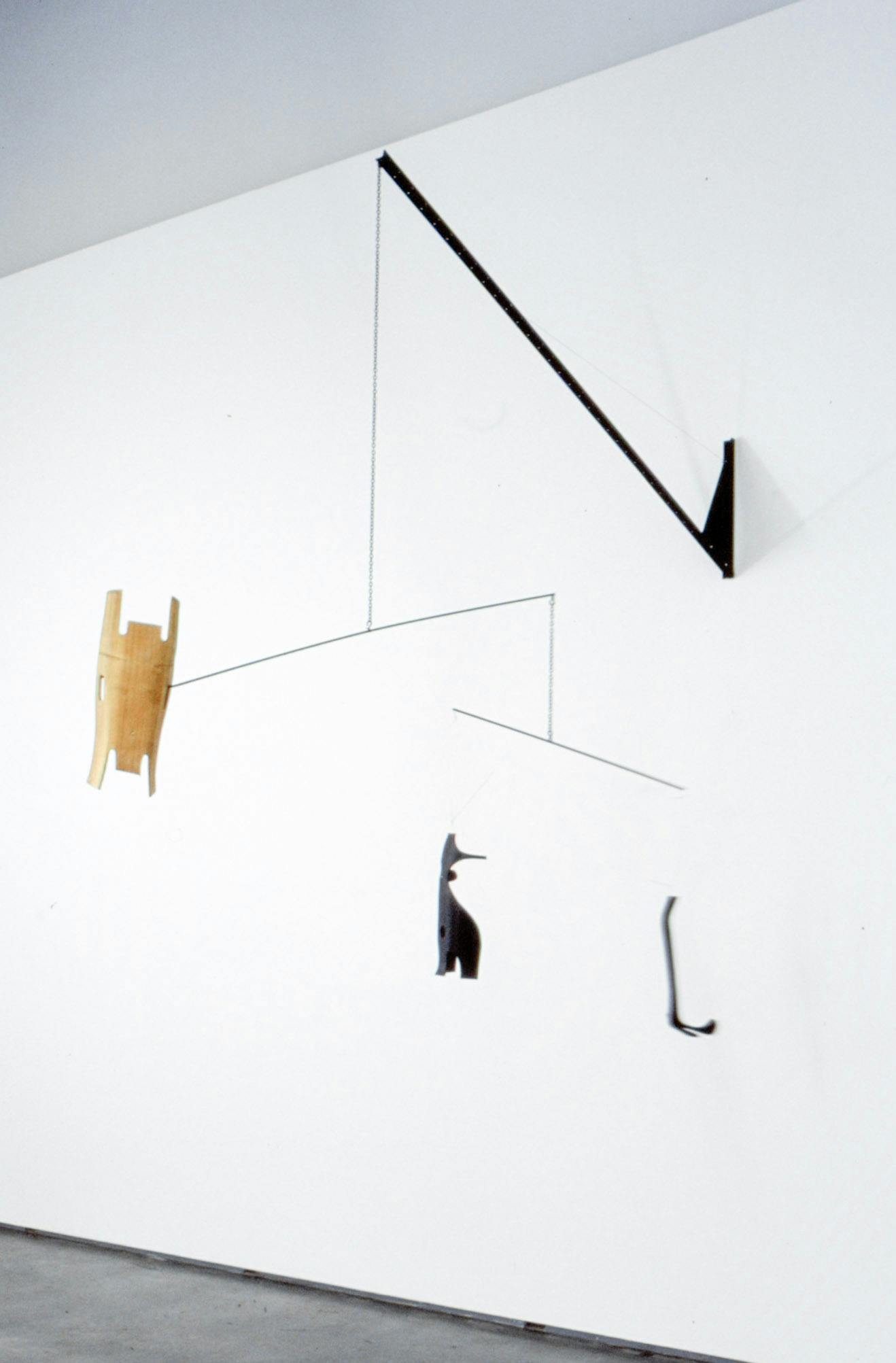 A sculpture is hanging from the wall in a gallery space. From the edge of the black stick attached to the wall, an object-attached wire is hanging down to the air. 