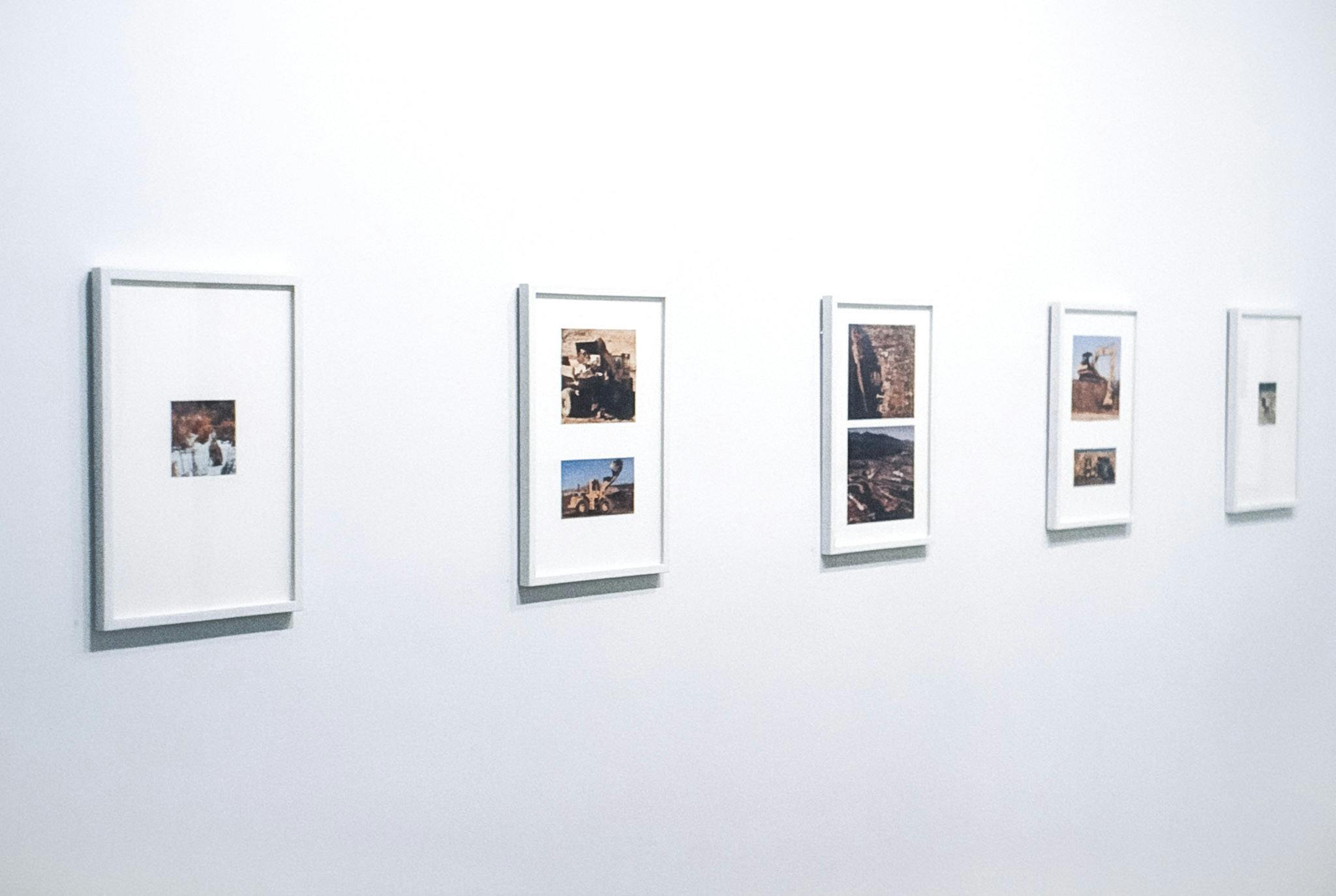 5 vertical, framed photographic works on a white wall. The three frames in the centre contain 2 photos each, all of construction sites. The two on the ends each have one small photo of a landscape. 