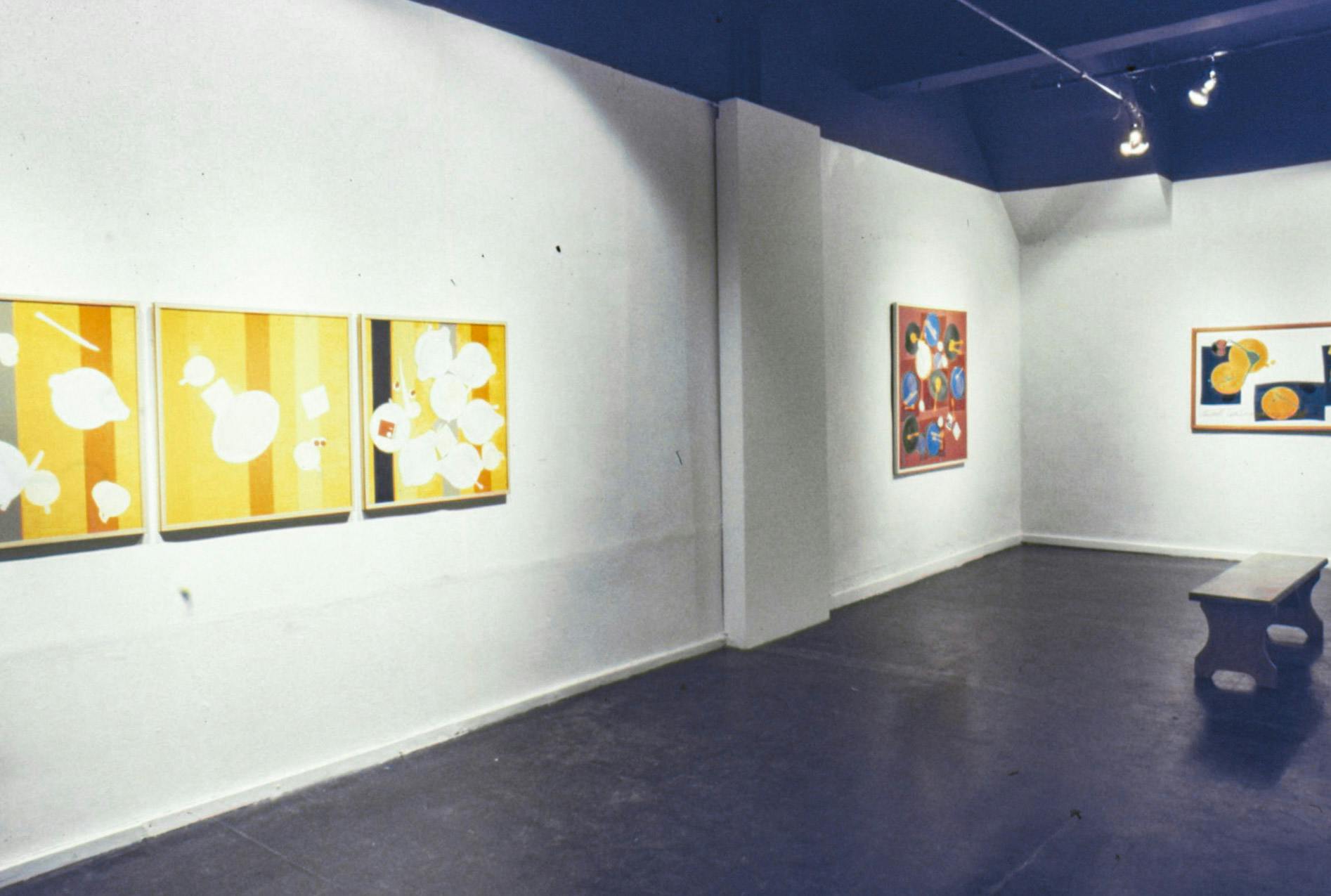 A gallery space with framed paintings on the walls and a bench on the floor. The paintings are colourful and show different dinnerware sets which have colourful patterns on the backgrounds.