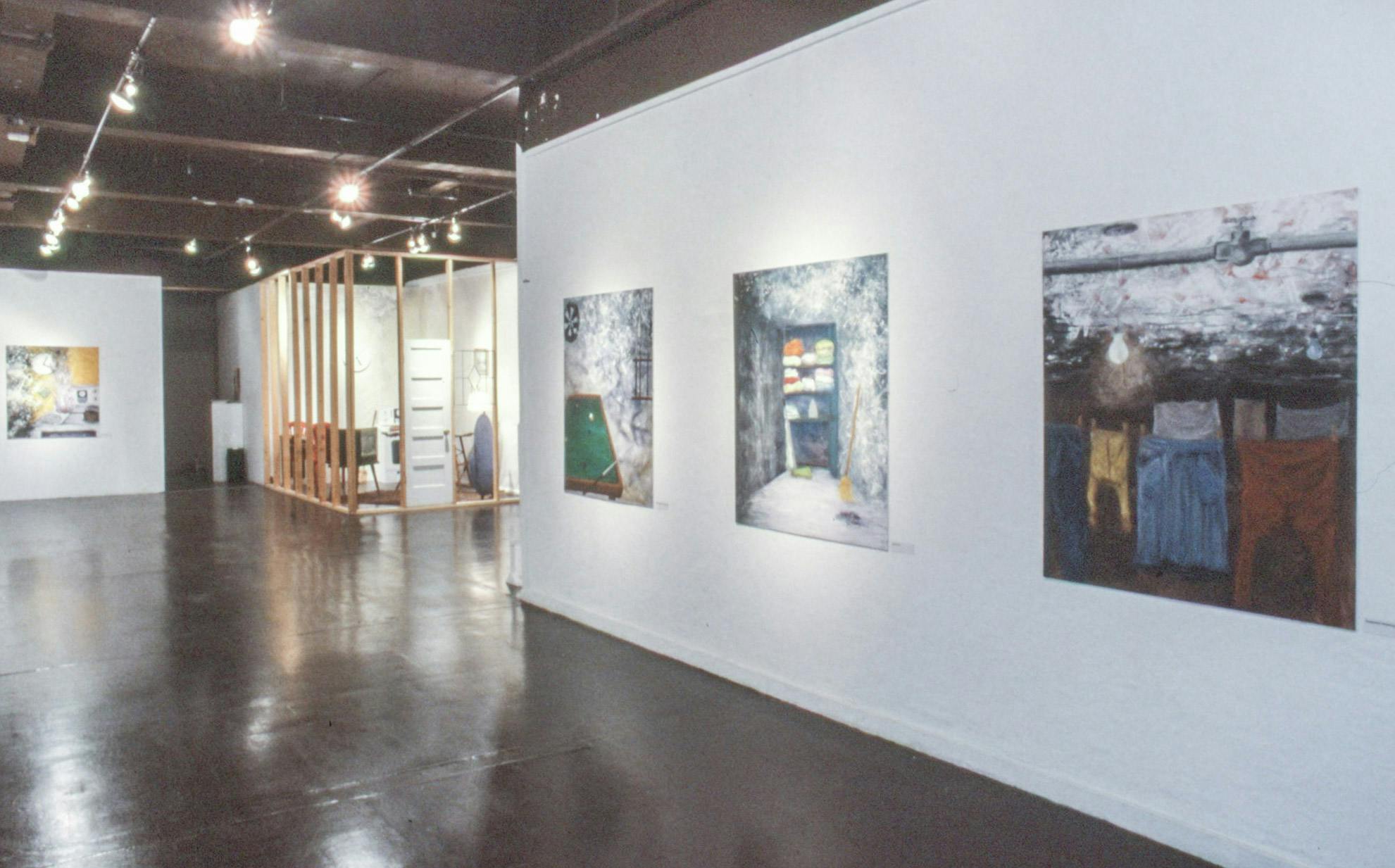 A gallery with paintings on the wall, and a large installation. The paintings show a pool table, a closet, and a clothesline in a basement. The installation is in a wooden frame with a white door.