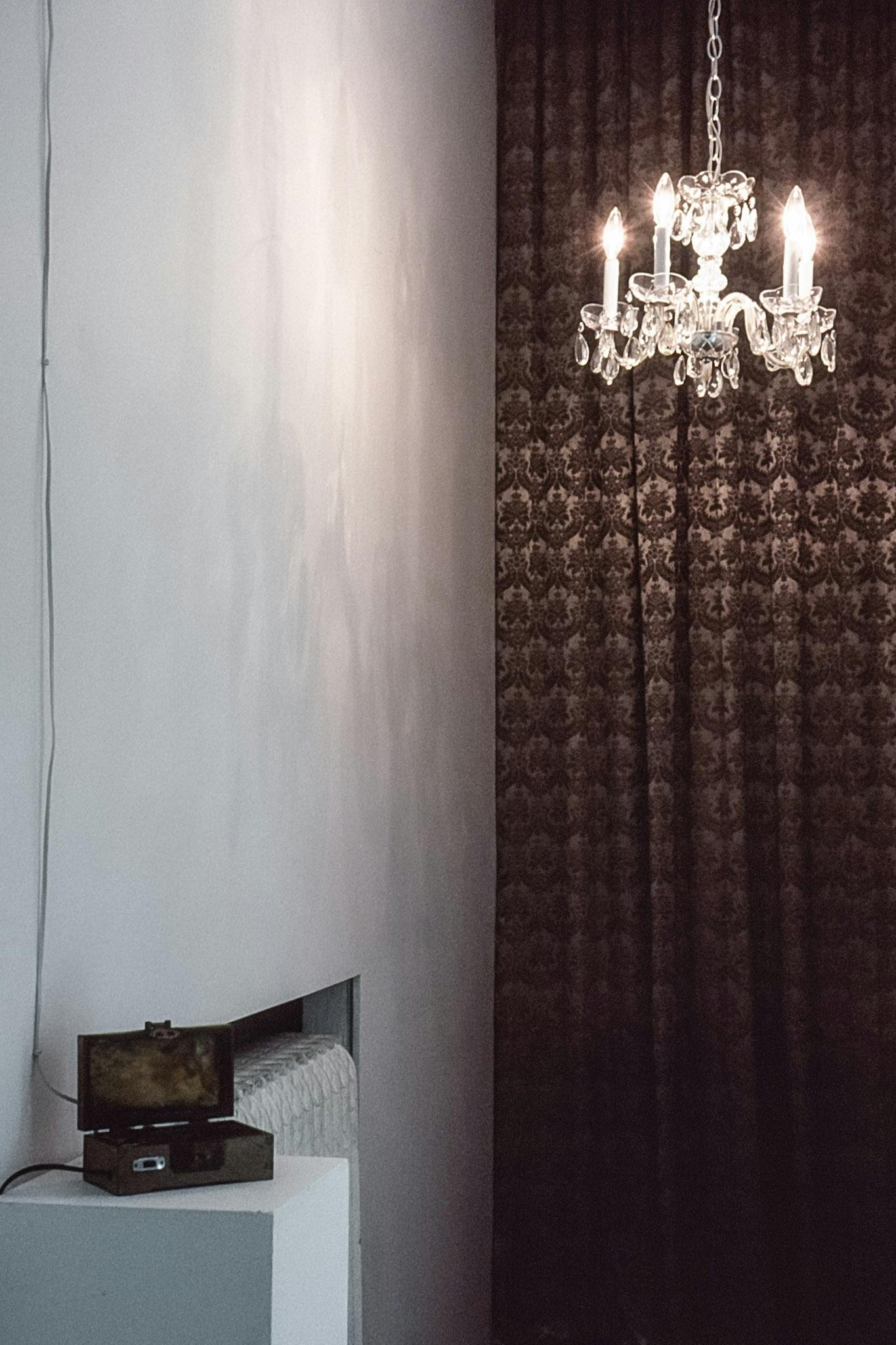 An installation image of a corner of a gallery space. A curtain is installed on a back wall. A small chandelier is hanging from the ceiling. A small box-like shaped object is installed on a pedestal. 