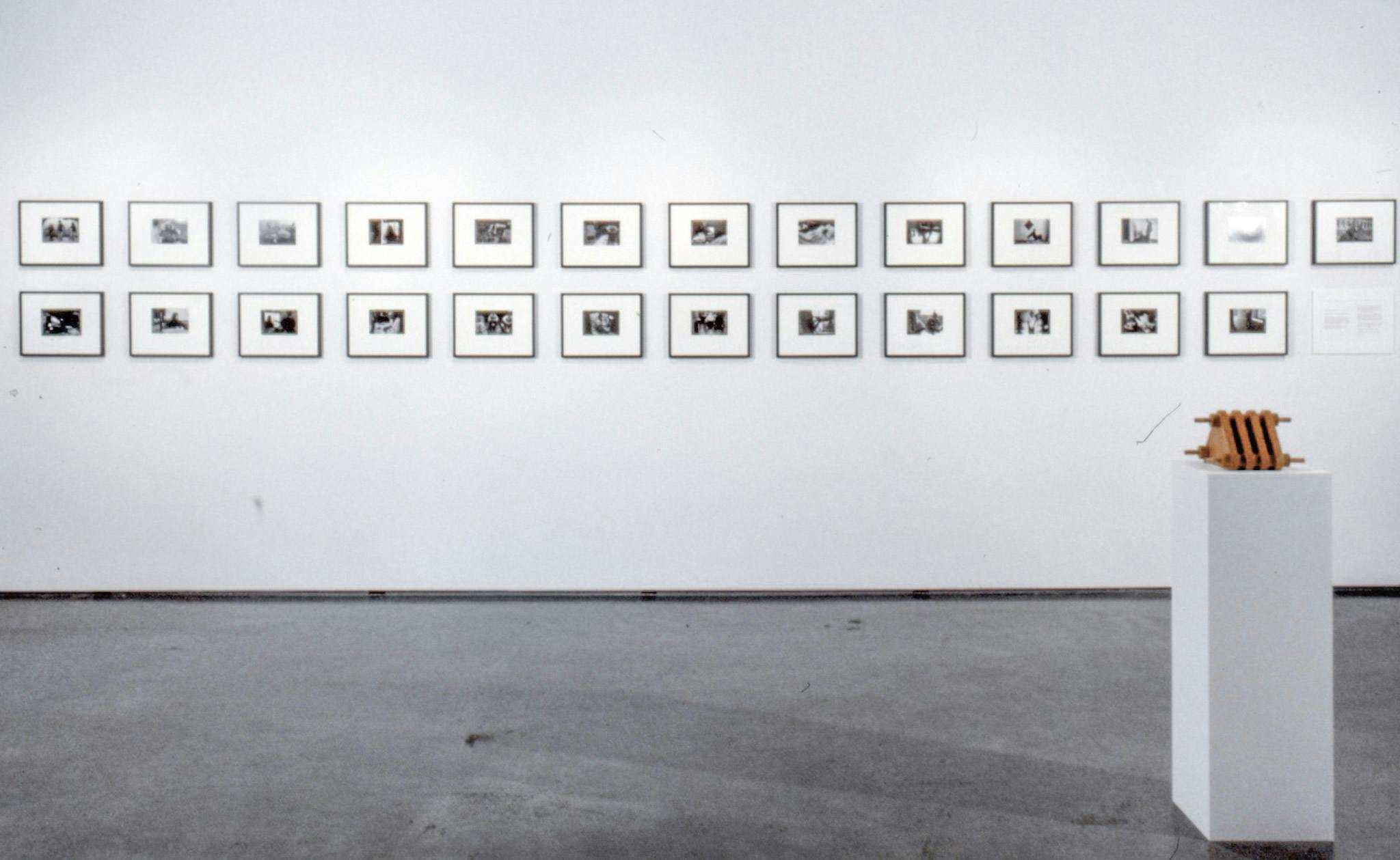 A number of black and whire photographs are mounted on the wall. They are arranged in two rows running horizontally to the floor. A wood sculpture on a pedestal stands in front of this wall.