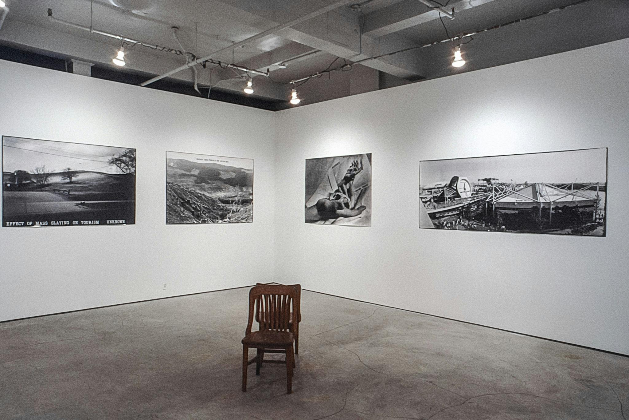 In the corner of a gallery, large black and white photos line the walls. The photos are horizontal and show landscapes and a set of hands. In the centre of the room, 2 wooden chairs sit back to back.