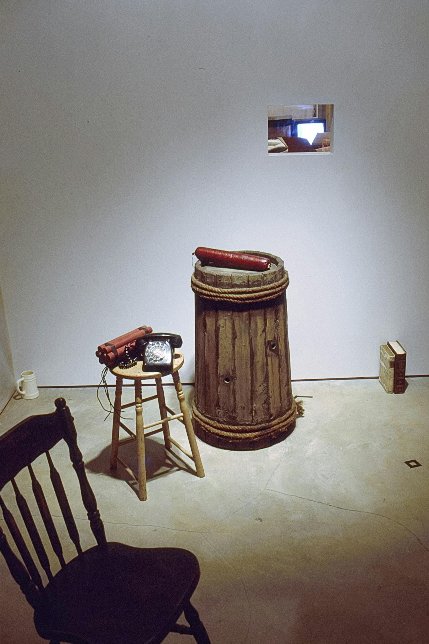 A wooden skinny barrel, stool, chair, two books, and a ceramic coffee mug are placed on the gallery floor. A black dial phone and a dynamite bundle are placed on top of the stool. 