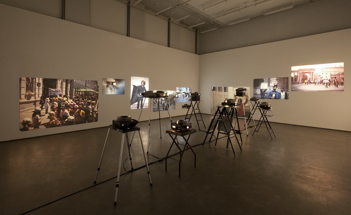 A dozen projectors are installed in the middle of a gallery space. They project photographs onto the walls, which are the documentations of Sharon Hayes’ public performances in various locations.