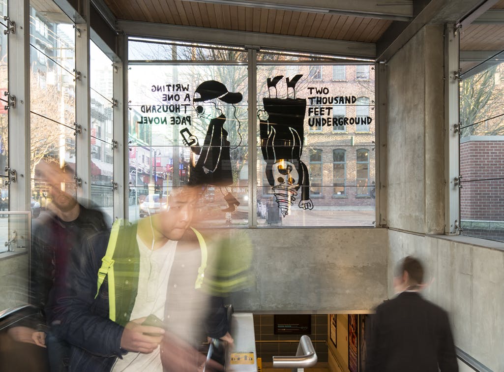 A graphic illustration of two figures printed in black vinyl on the glass face of a train station. One is walking and the other is upside down with a drill-like head. Bold text is on either side.  
