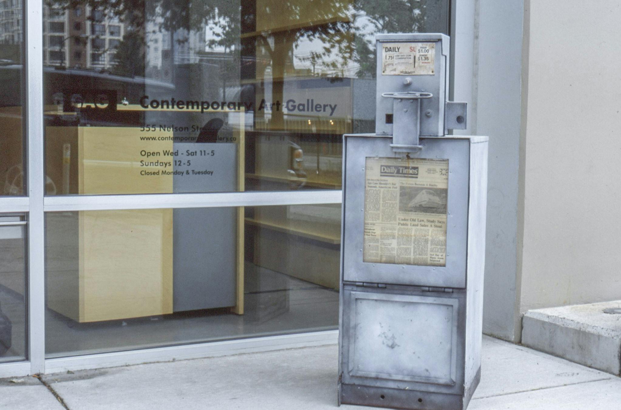 A metal made newspaper vending machine sits outside of the CAG’s glass entrance. This silver coloured machine looks old. It sells Daily Times.