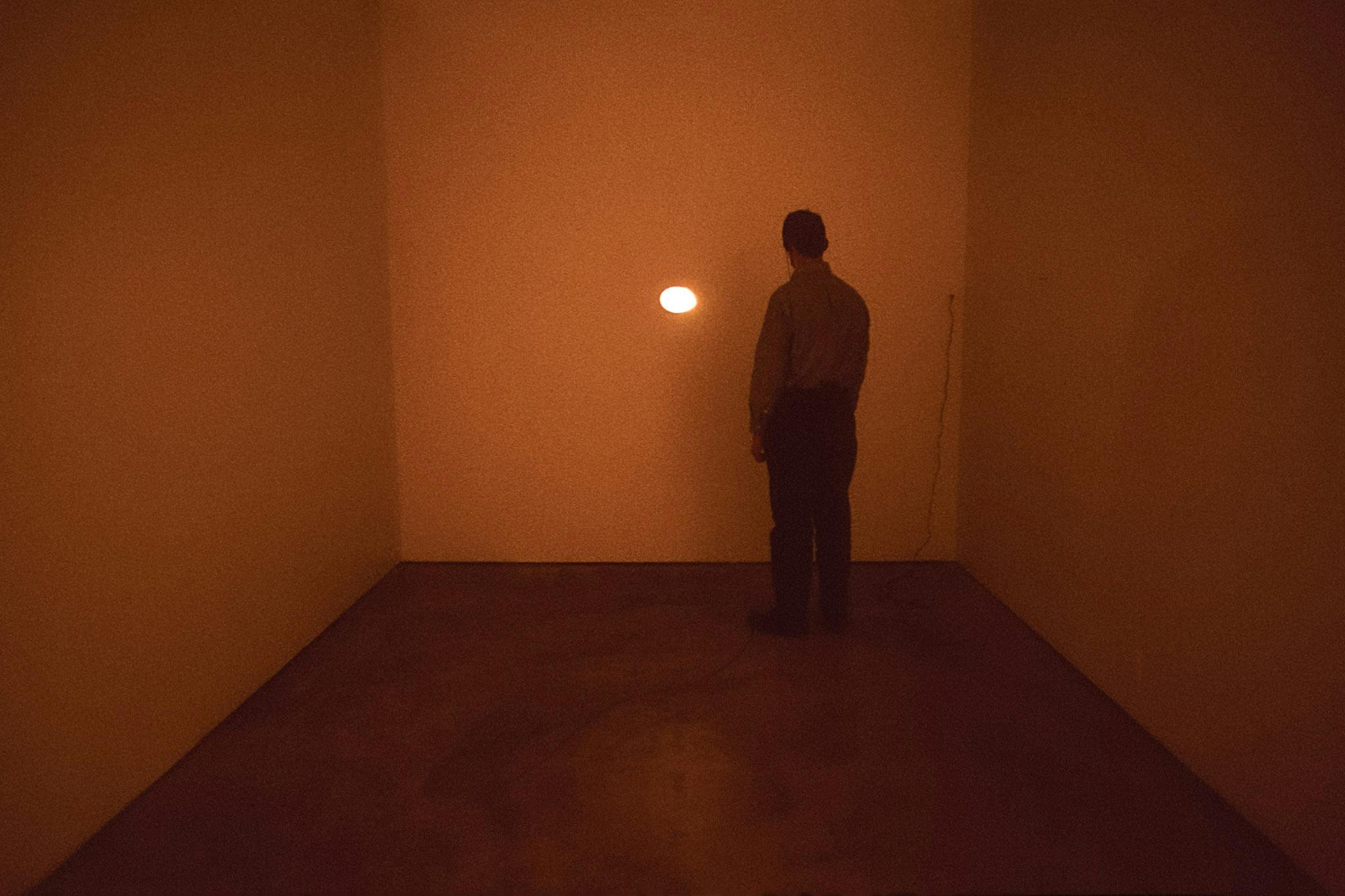 A man is looking at the artwork attached to the white gallery wall. The piece has a small oval shape and it glows orange light in a darkened gallery space. 