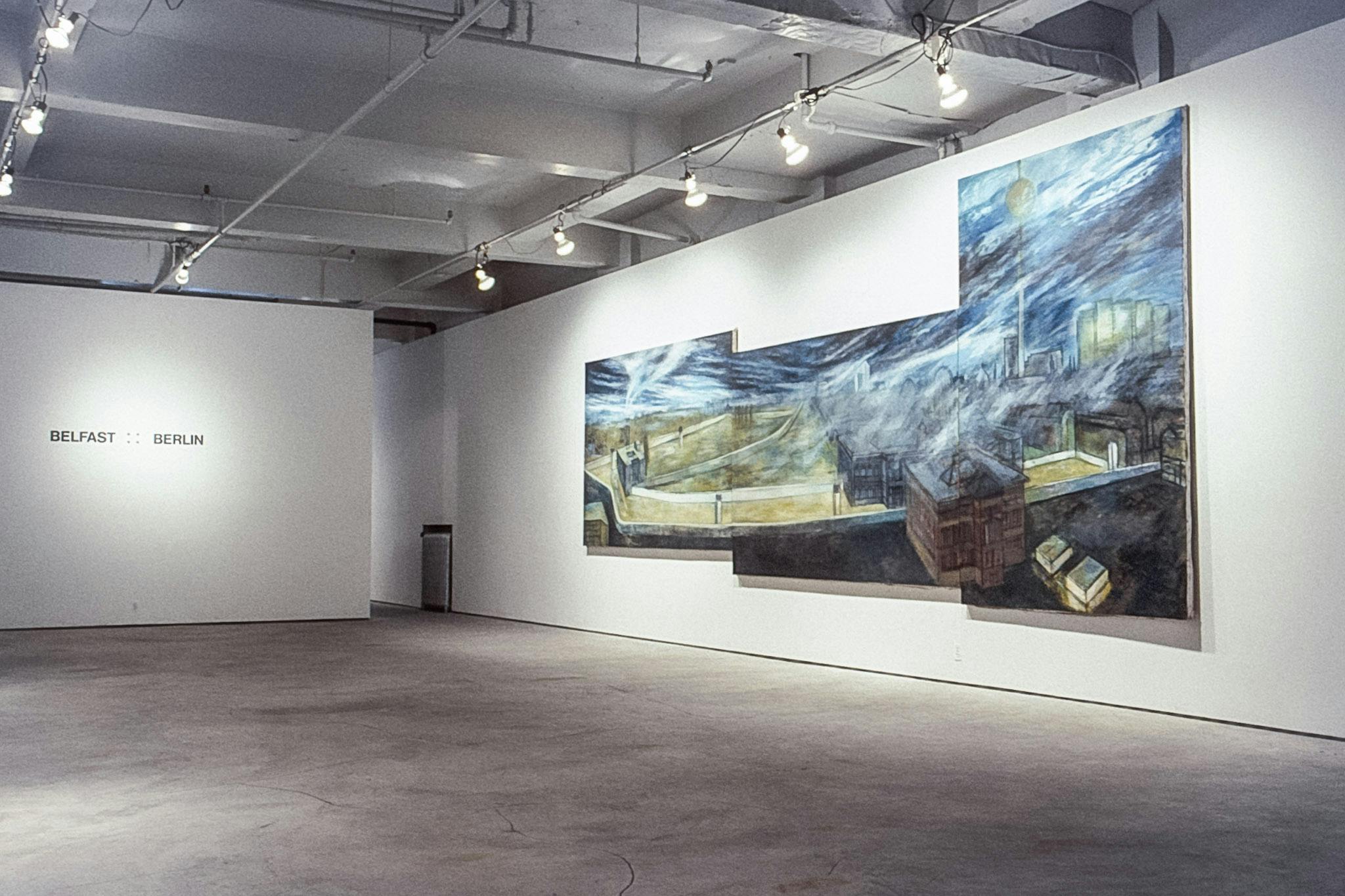 A gallery space showing a large blue and yellow painting made up of 3 canvases mounted on a wall. On another wall, there is large black text that reads "Belfast : : Berlin."