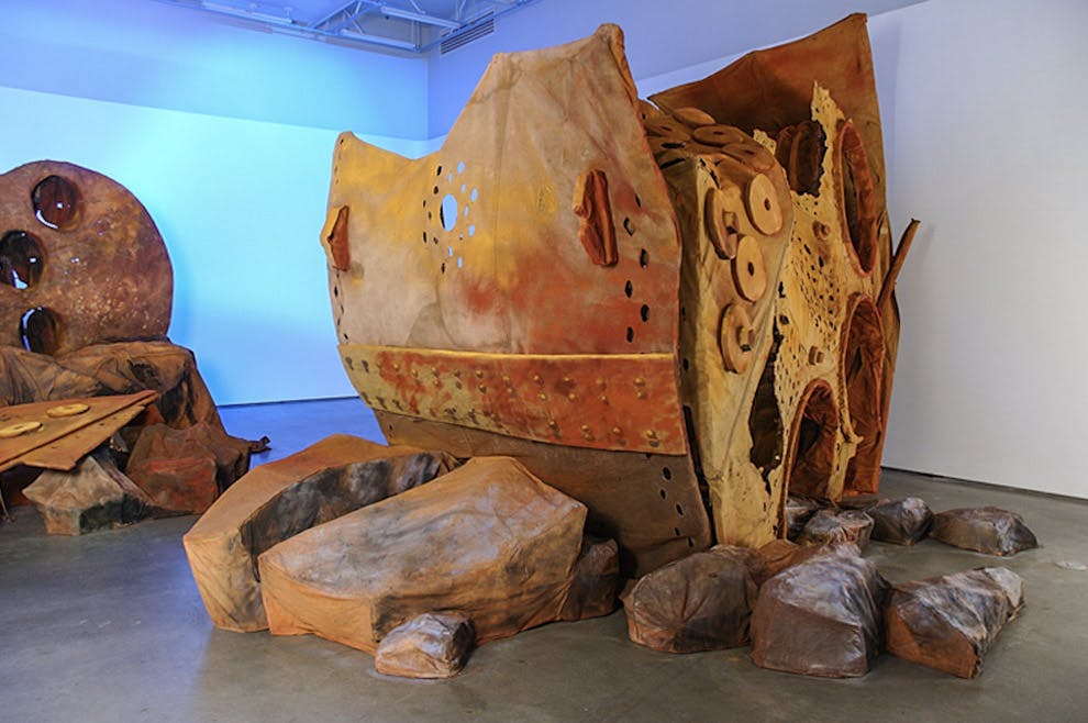 An installation image of large artworks by Donald Lawrence inside a gallery. An assemblage of distorted and decayed metal parts of some large industrial machine is placed on the floor. 