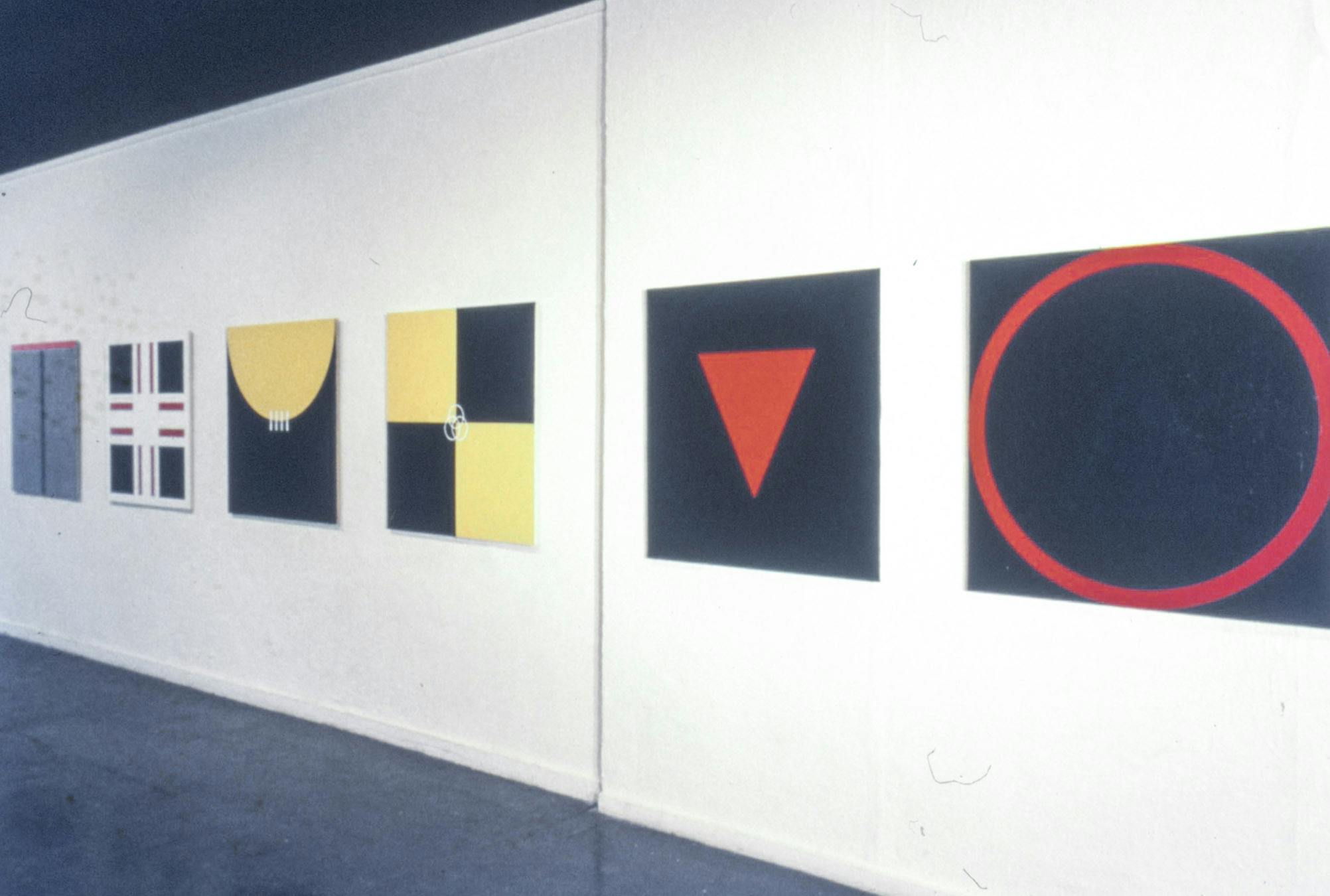 Six large paintings on a wall. They are all on square canvases and show different geometric shapes including circles, squares, triangles, and crosses. They are red, black, white, grey, and yellow.