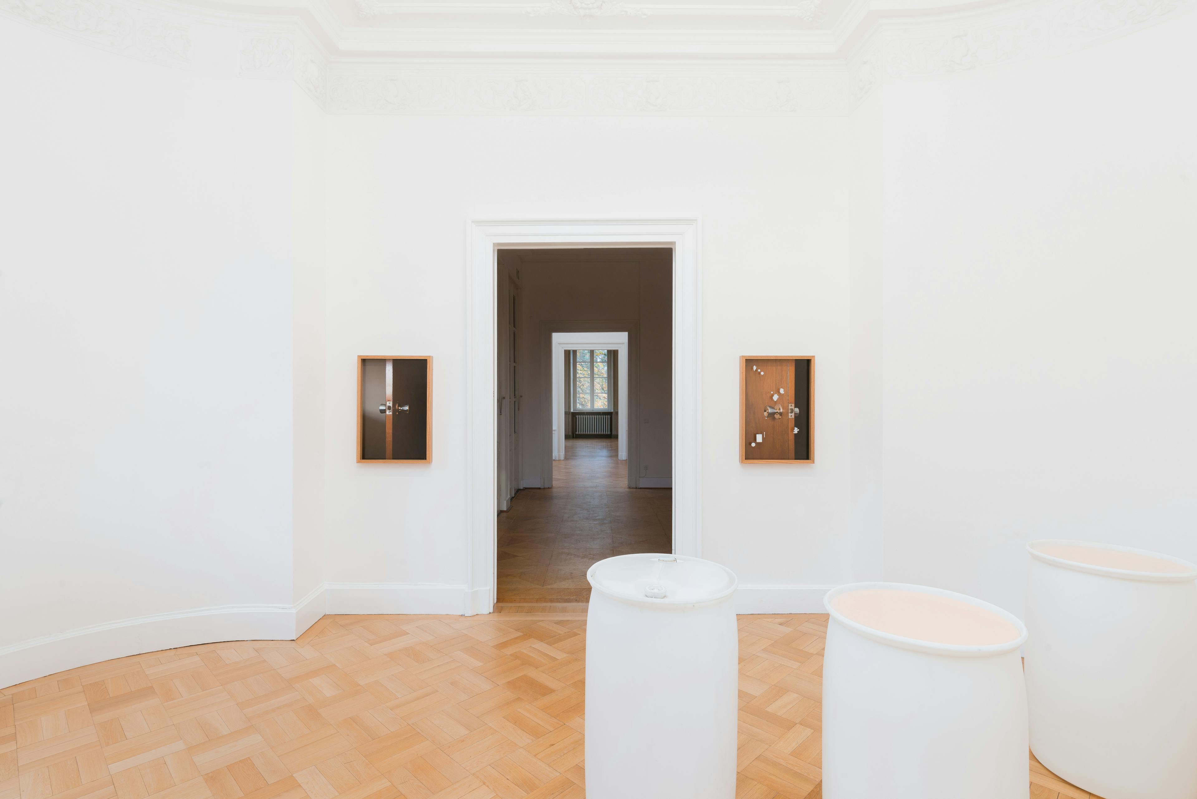 Several artworks are installed in a gallery space. Three white plastic barrel sculptures are placed on the wood tile floor. Two wood-framed photographs of a doorknob are mounted on the white wall