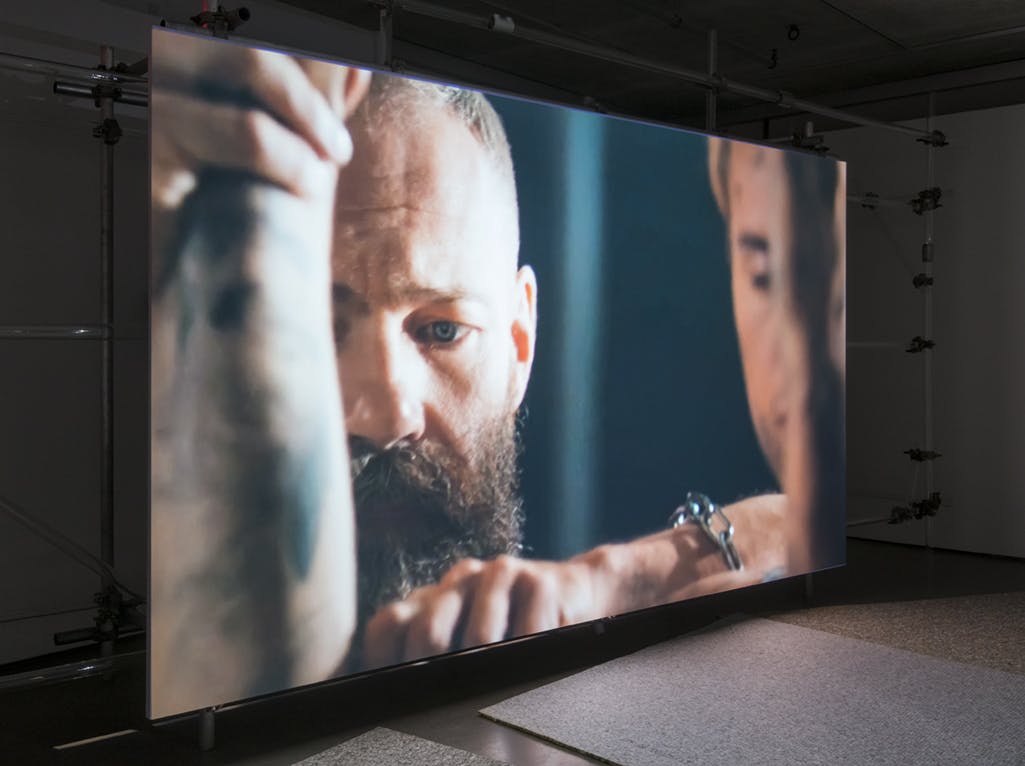 A video work by Patrick Staff is projected onto a large screen in a darkened gallery. The camera is closely focused on a bearded person’s face, who stands behind other figures.