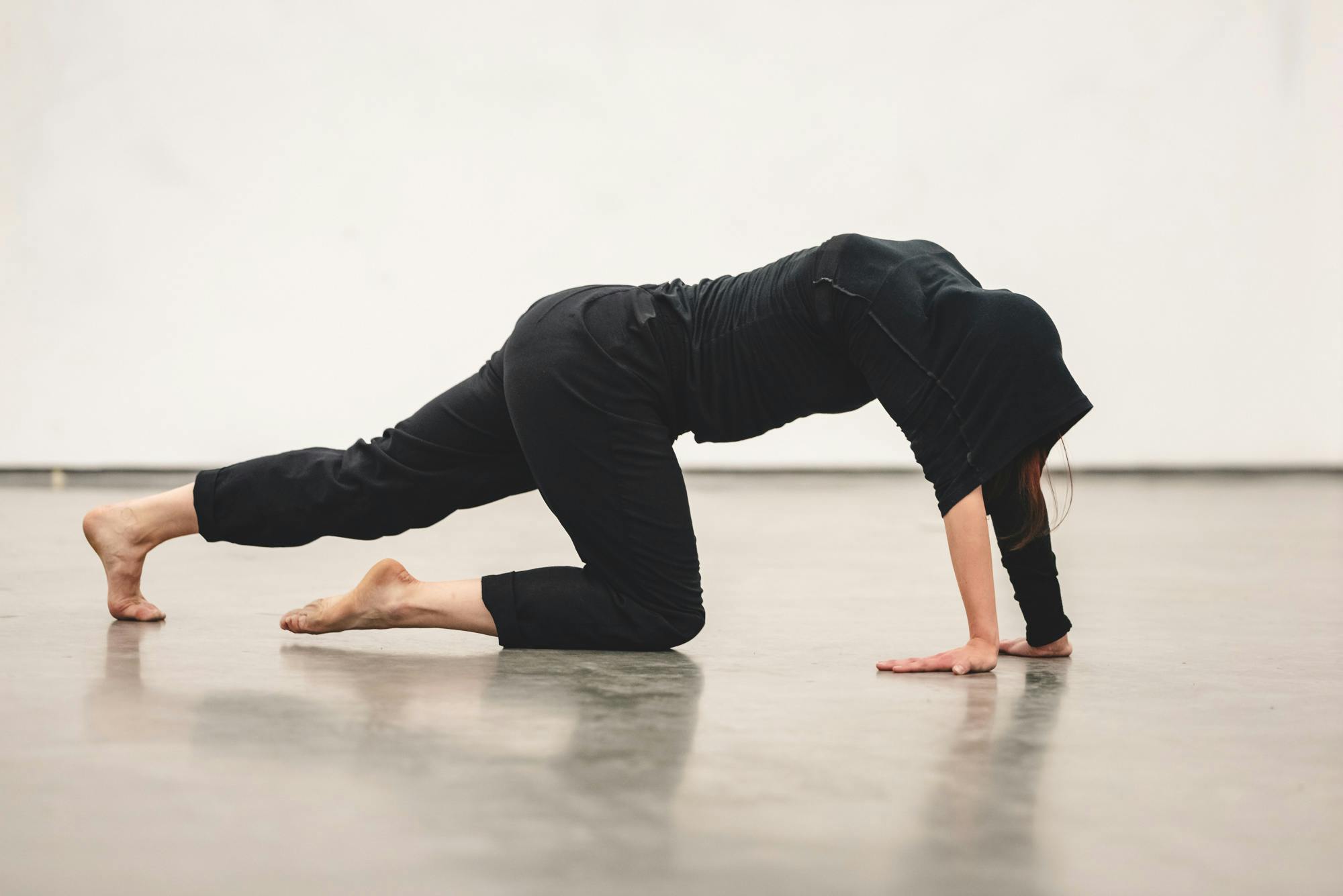 A photograph of a person in a black outfit performing in the gallery. The person stretches their arms on the floor and kneels on their right knee, stretching the other leg. 