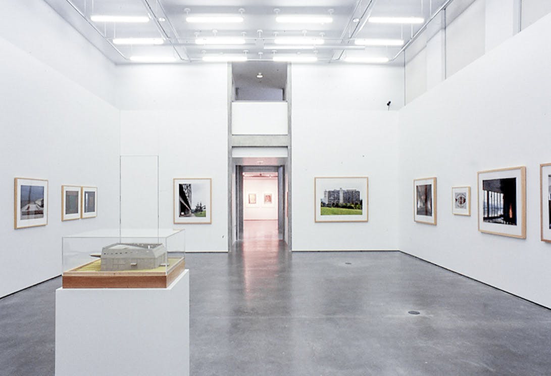 A large gallery space with a large entryway that is brightly lit. On the walls there are several framed photos of architectural spaces. ON the floor, there is a plinth holding a small model building. 