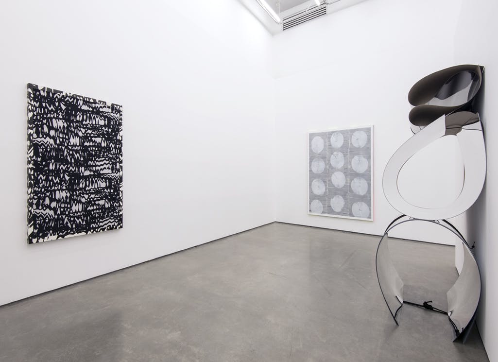 Two abstract, black and white paintings and one sculpture are installed in a gallery space. The sculpture is made of large, arched and bent sheets of reflective material stacked atop one another. 