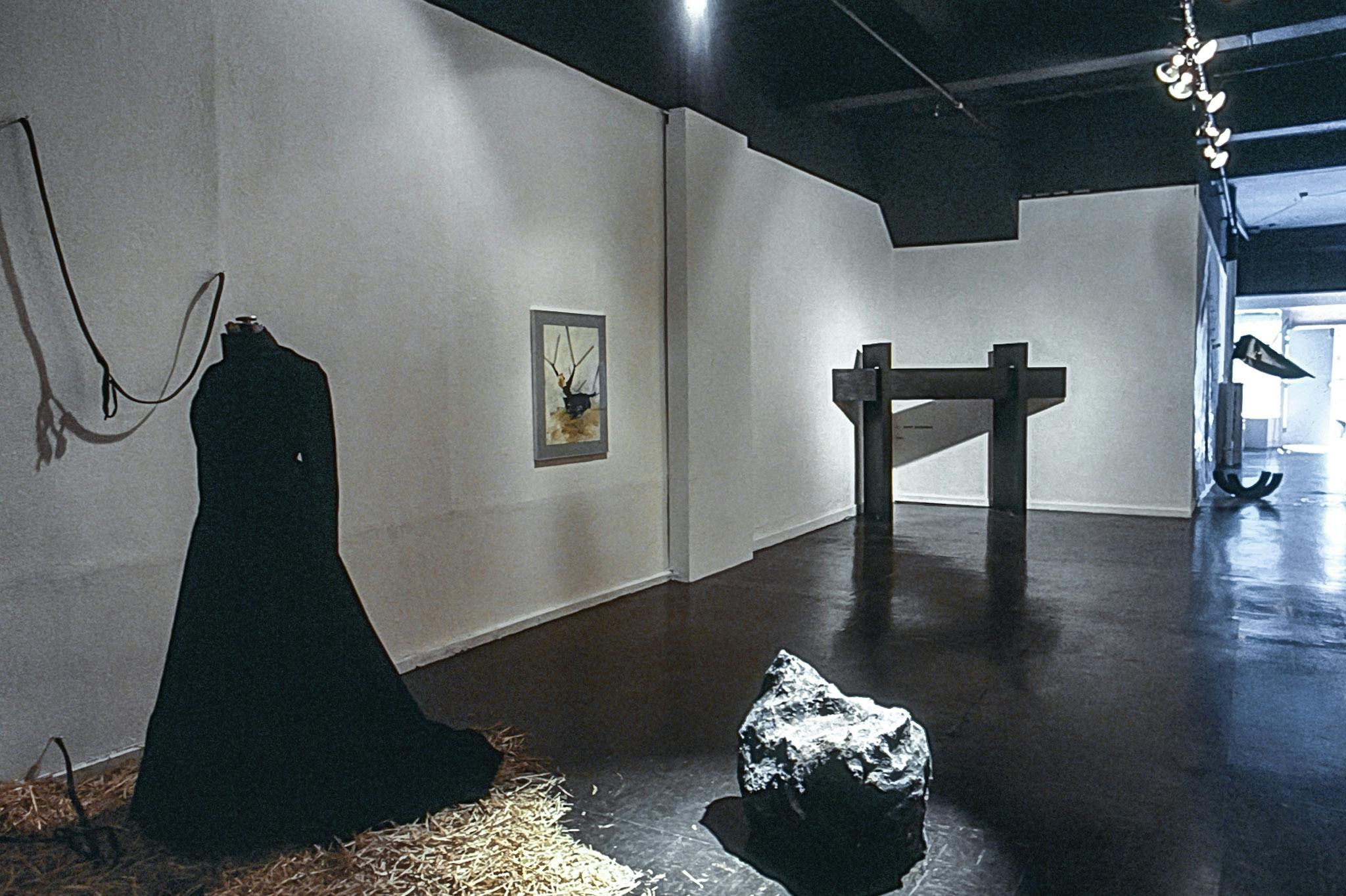 Several artworks in the corner of a gallery. The works include a black dress standing on hay, a rock, a small painting, and a small, black wooden arch with text on the wall behind it.