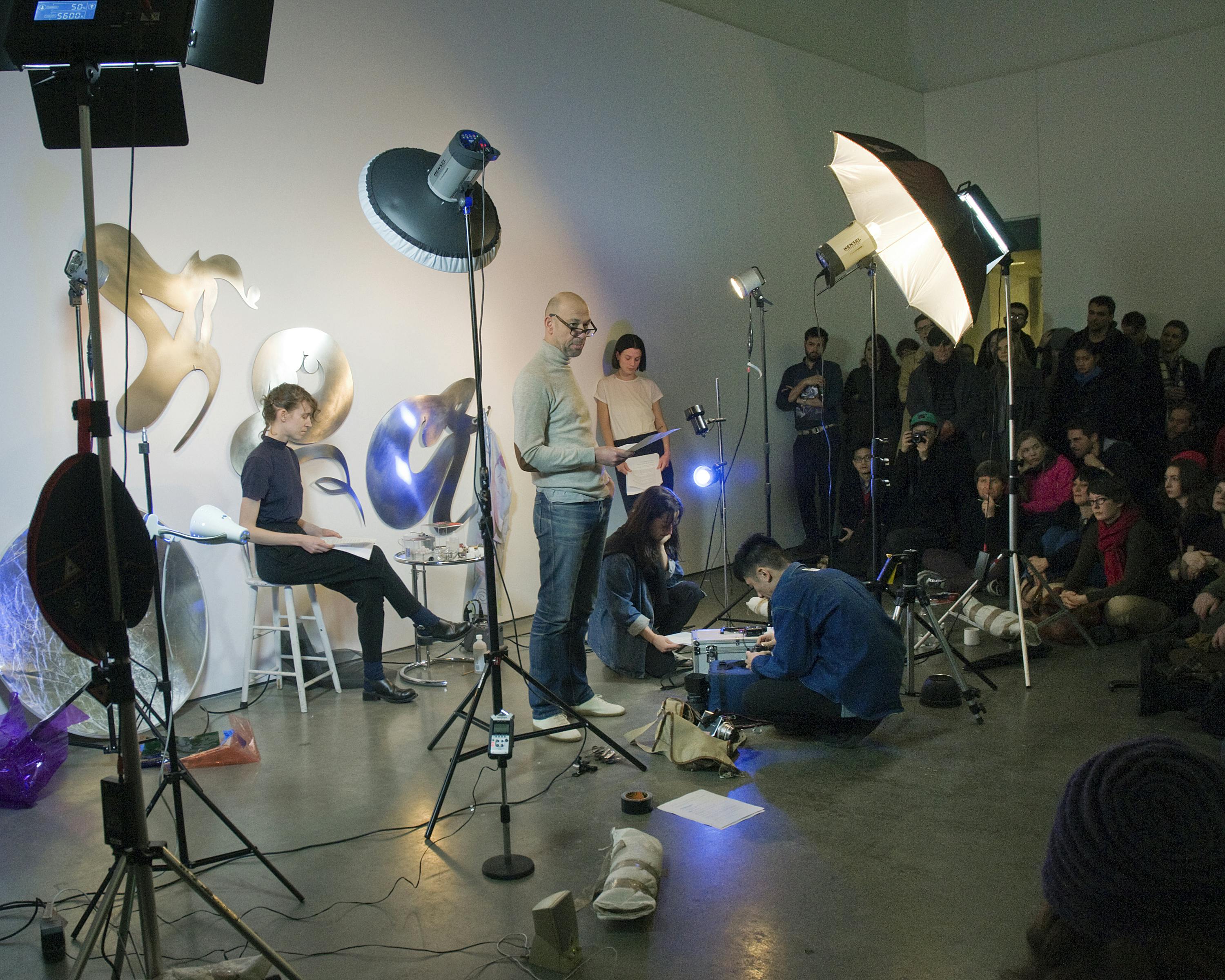 Five people are posing in a small photo booth set in front of a wall in a gallery. A person standing in the middle of the booth looks at the audience sitting around the set.