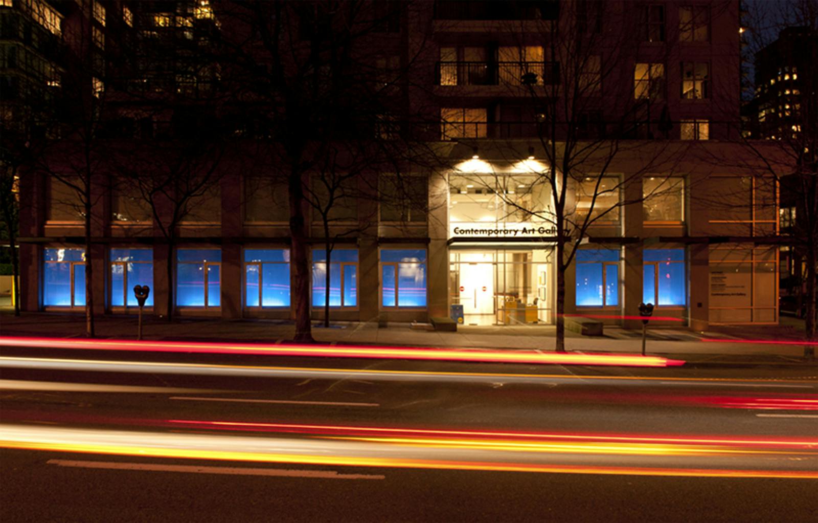 Scott Massey’s works are installed across CAG’s facade windows. These windows are blue with white light glowing at the bottom. It is nighttime, and yellow lights are glowing from the other buildings.