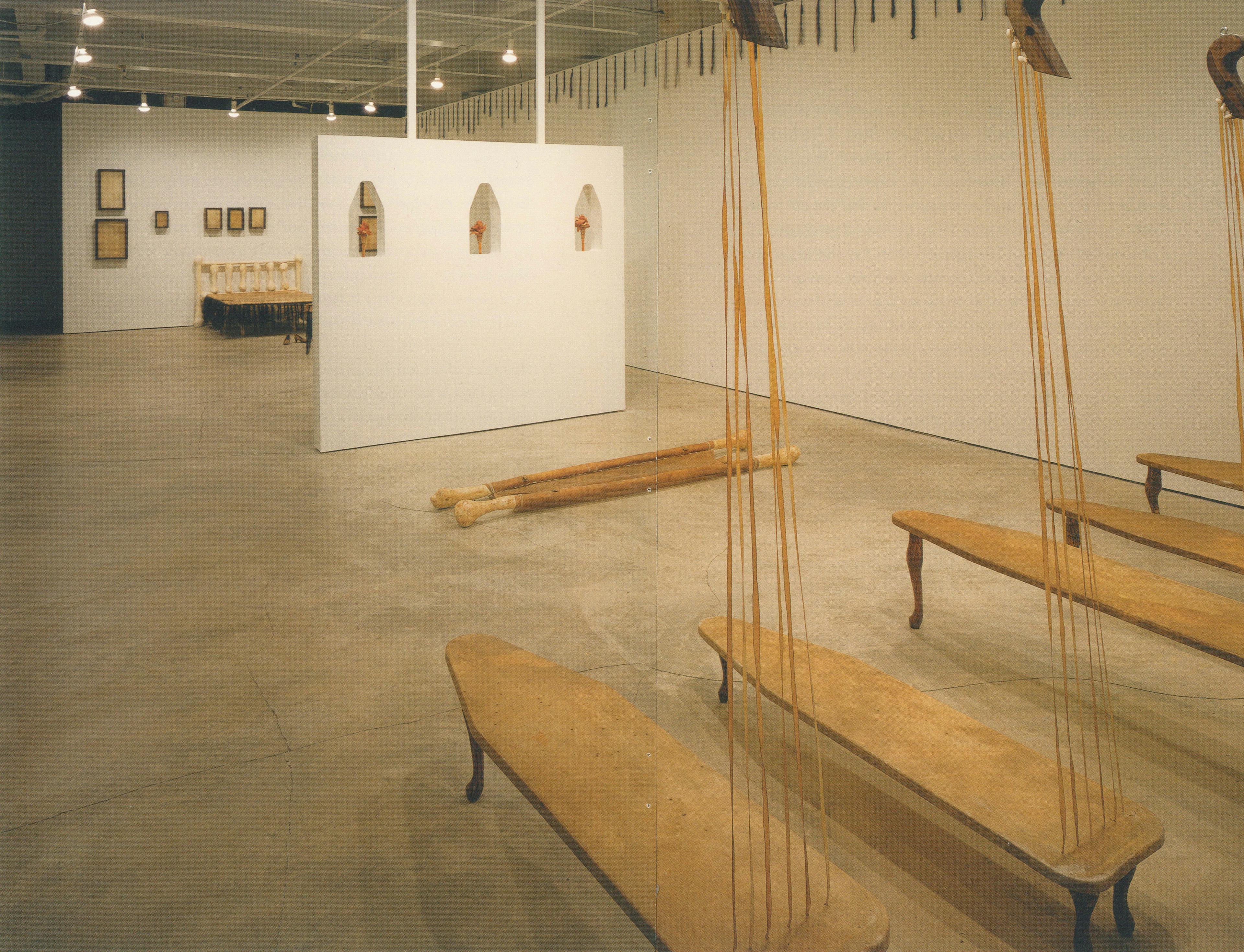 Various artworks are installed in a gallery space. Five low wooden benches, or ironing boards, are placed in a row. A pair of long wooden poles are laid on the floor some feet away from the benches. 