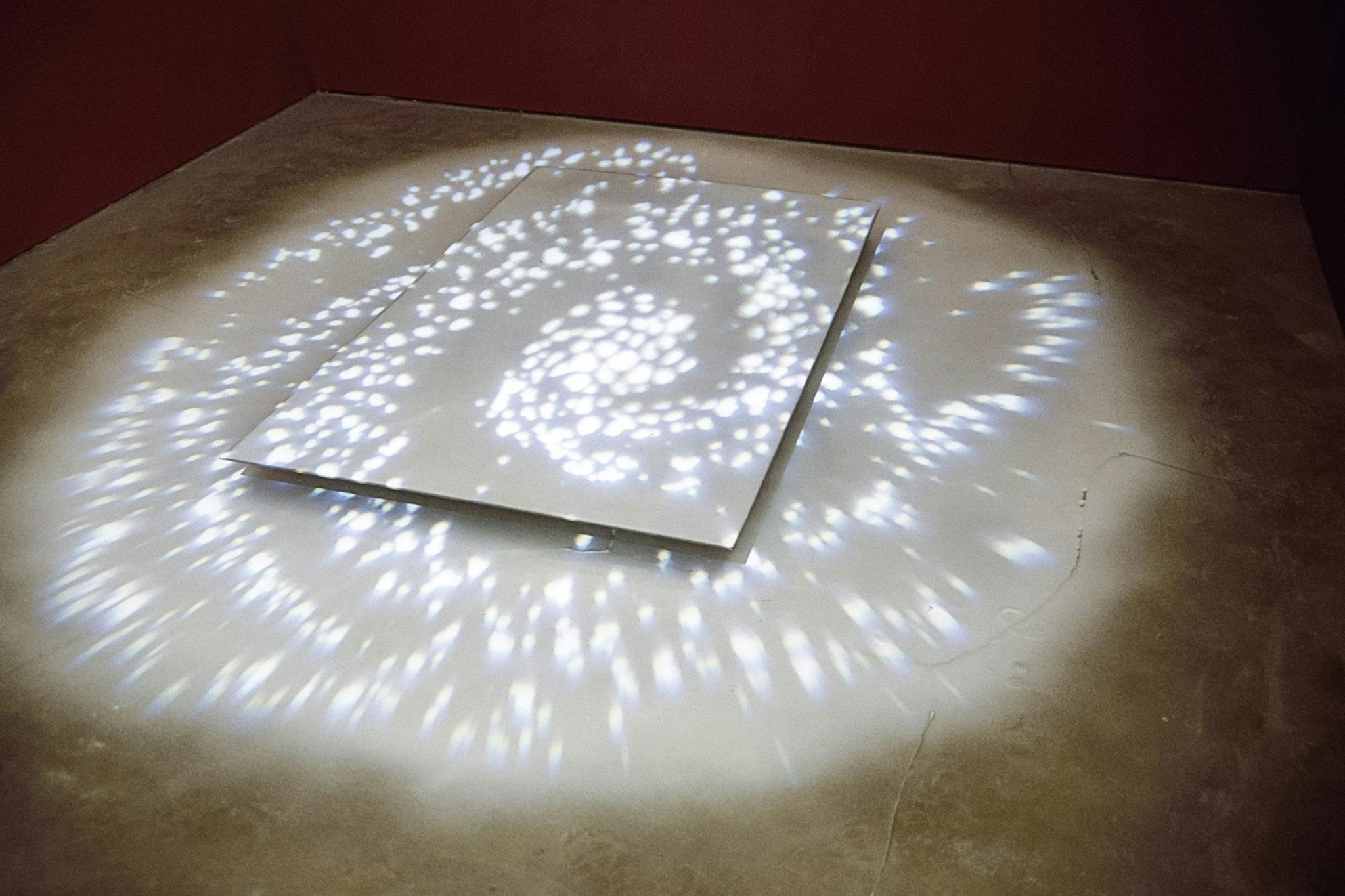 An installation on the white gallery floor consists of a white panel and a projection from the ceiling. A white spiral shape is projected over the panel. The gallery walls are painted in red. 