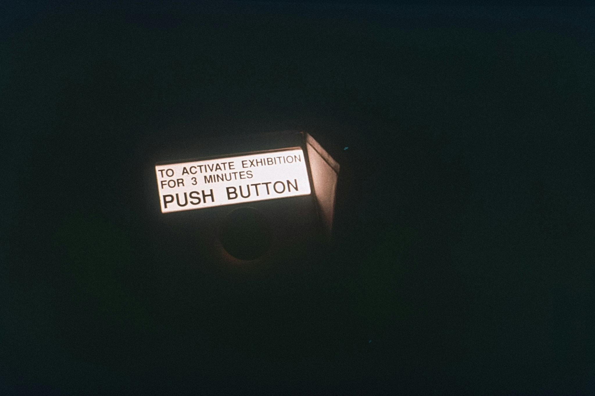 A darkened entrance to a gallery space, with a small bright sign glowing above a round buzzer button. The sign reads "to activate the exhibition for 3 minutes, push button."  
