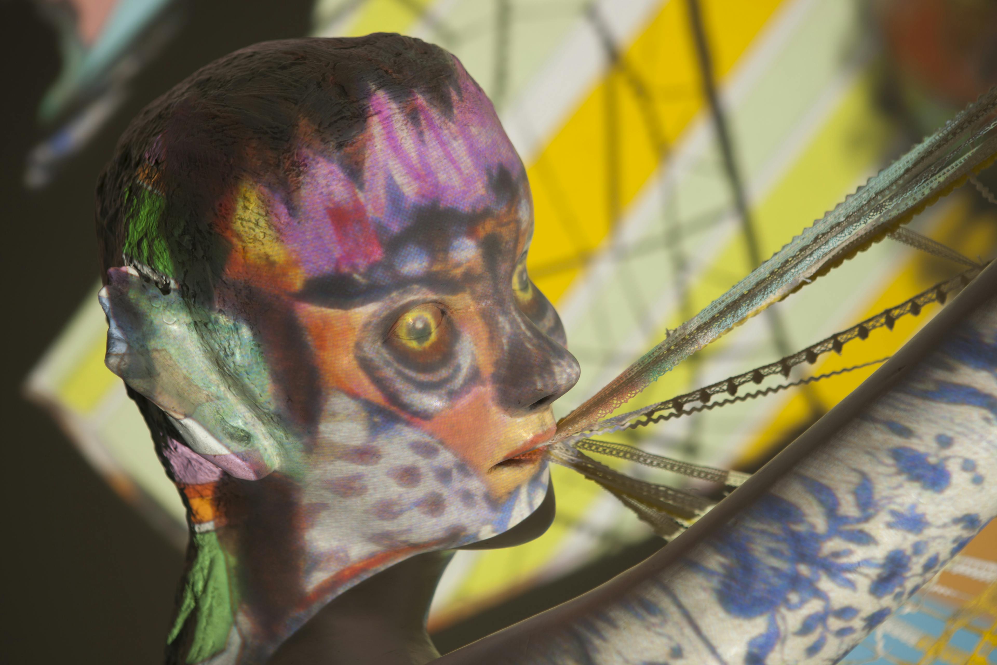 A human head-like plaster sculpture lit by a colourful, patterned projection. Owl eyes in the projection line up with those of the sculpture’s and lace strings emerge from its mouth.