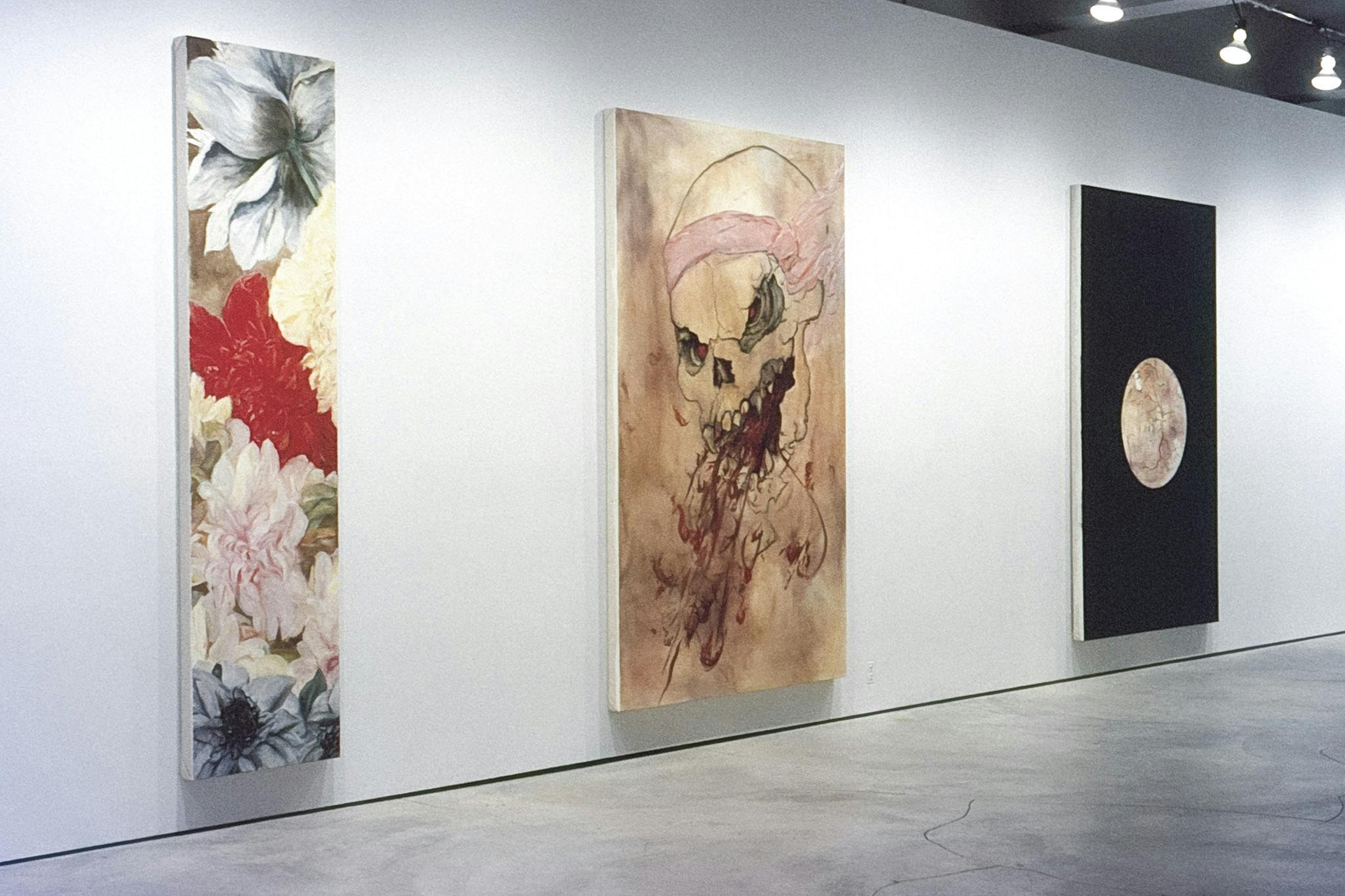 Three vertical paintings in a gallery. One has several large flowers layered together, one has a skull spewing blood from its mouth, and one is black with a beige circle that has a cracked texture.