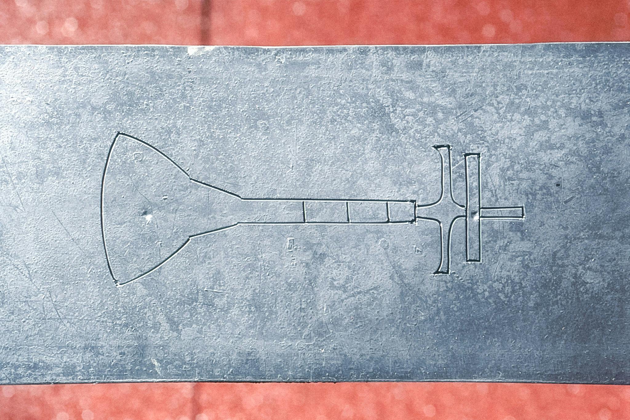 A two-dimensional image of a manual air pump is carved on a metal surface. The image of the air pump is horizontal, with the bottom of the air pump on the left, and the top of it on the right. 