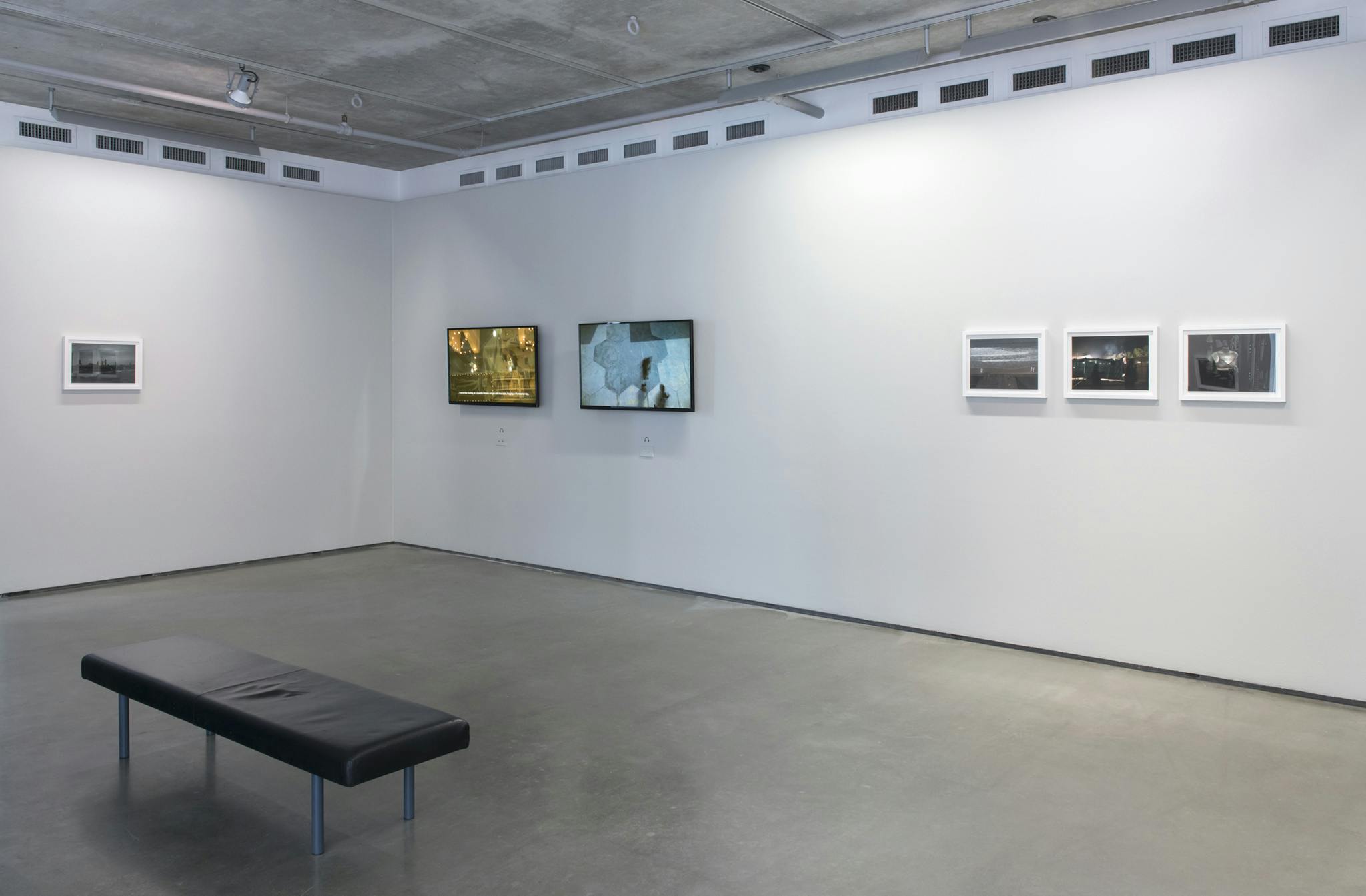 Four framed photographs and two monitors hang on two walls. The left wall has one photograph and the right wall has three photographs and two monitors. One bench sits in the middle of the room.