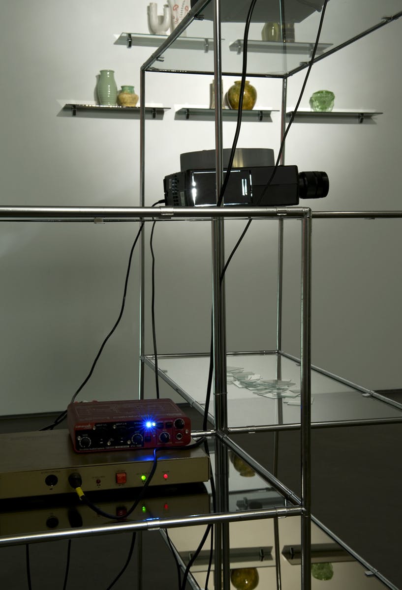 Detail image of glass shelves installed in the middle of a gallery space. Two projectors are placed at various heights. A pile of glass slides for a microscope is placed on one shelf.