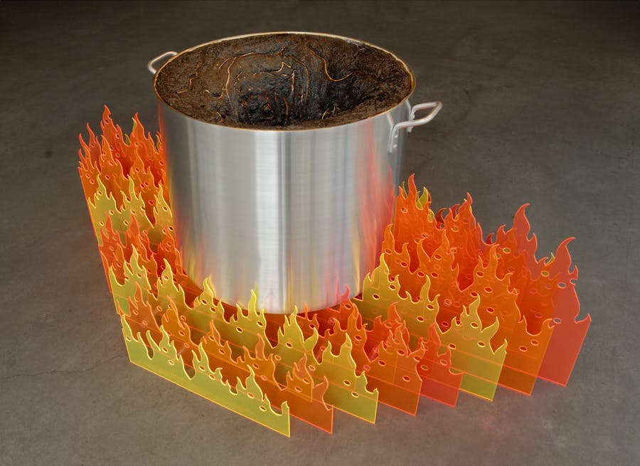 A silver stockpot is placed on a gallery floor. A dark brown form fills the inside of this pot. Orange and pink flame-shaped pieces surround the pot. 