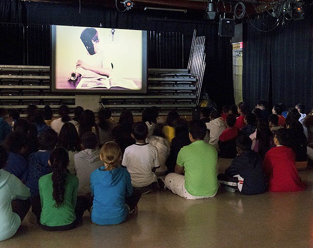 Primary school age children sit on the floor in a gym and watch a video screening. On the screen, a boy wearing a black mask is dipping a brush into ink with a thick photo book in front of him.