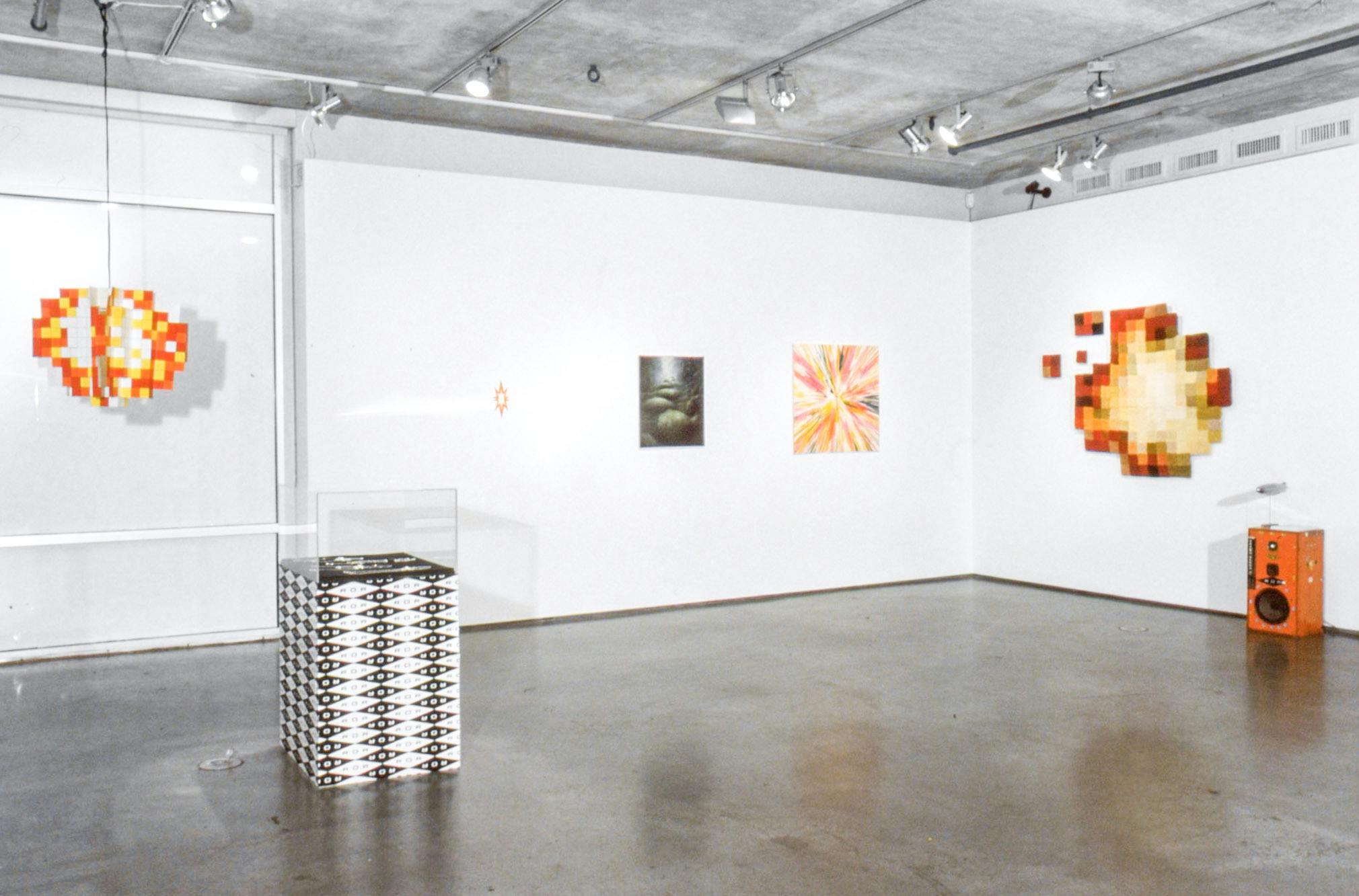 Installation image of works in a gallery. A display case sits in the middle of the floor. Three pictures are mounted on the far wall. An orange-brown sculpture is mounted on the other wall. 