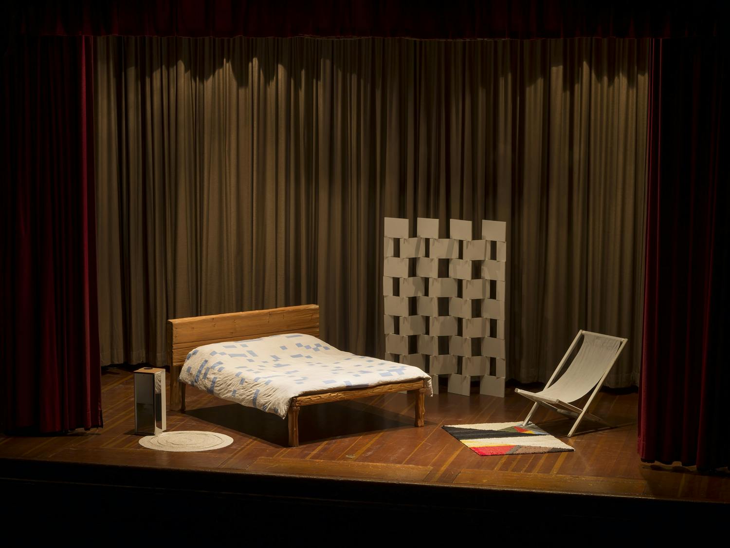 An image of a stage set framed by theatre curtains with brown curtains in the background. On stage is a set of a bedroom containing a bed, carpets, a small table, a room divider, and lounge chair.