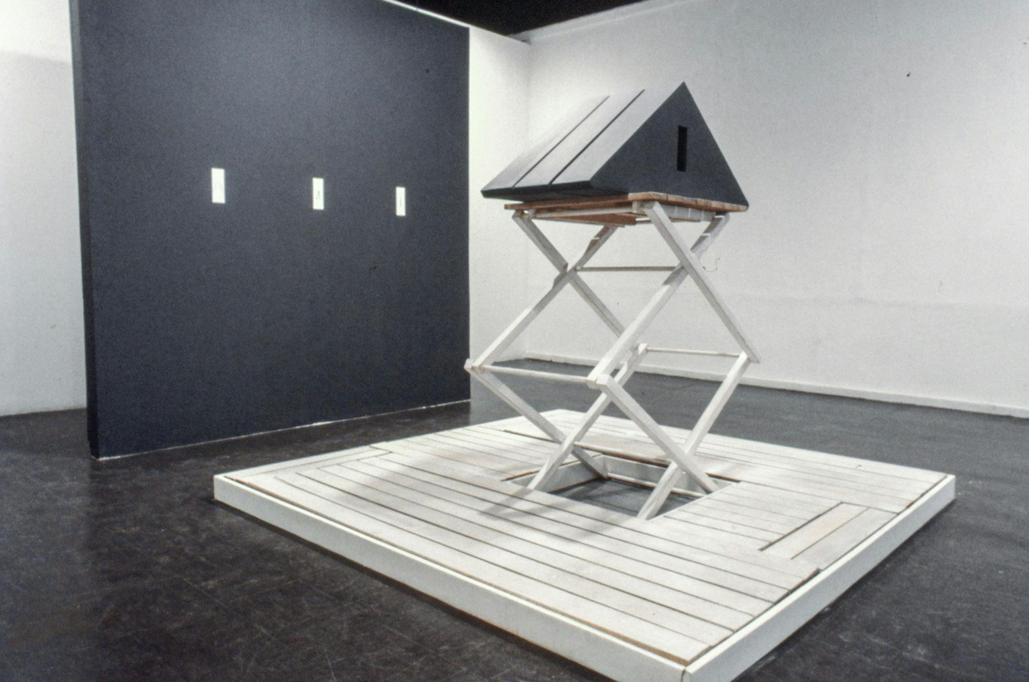 A closeup of a sculpture in a gallery. It is made of 3 narrow black pyramids stacked together on a scissor lift, placed on white wood planks. Behind the sculpture, there is a black divider wall.