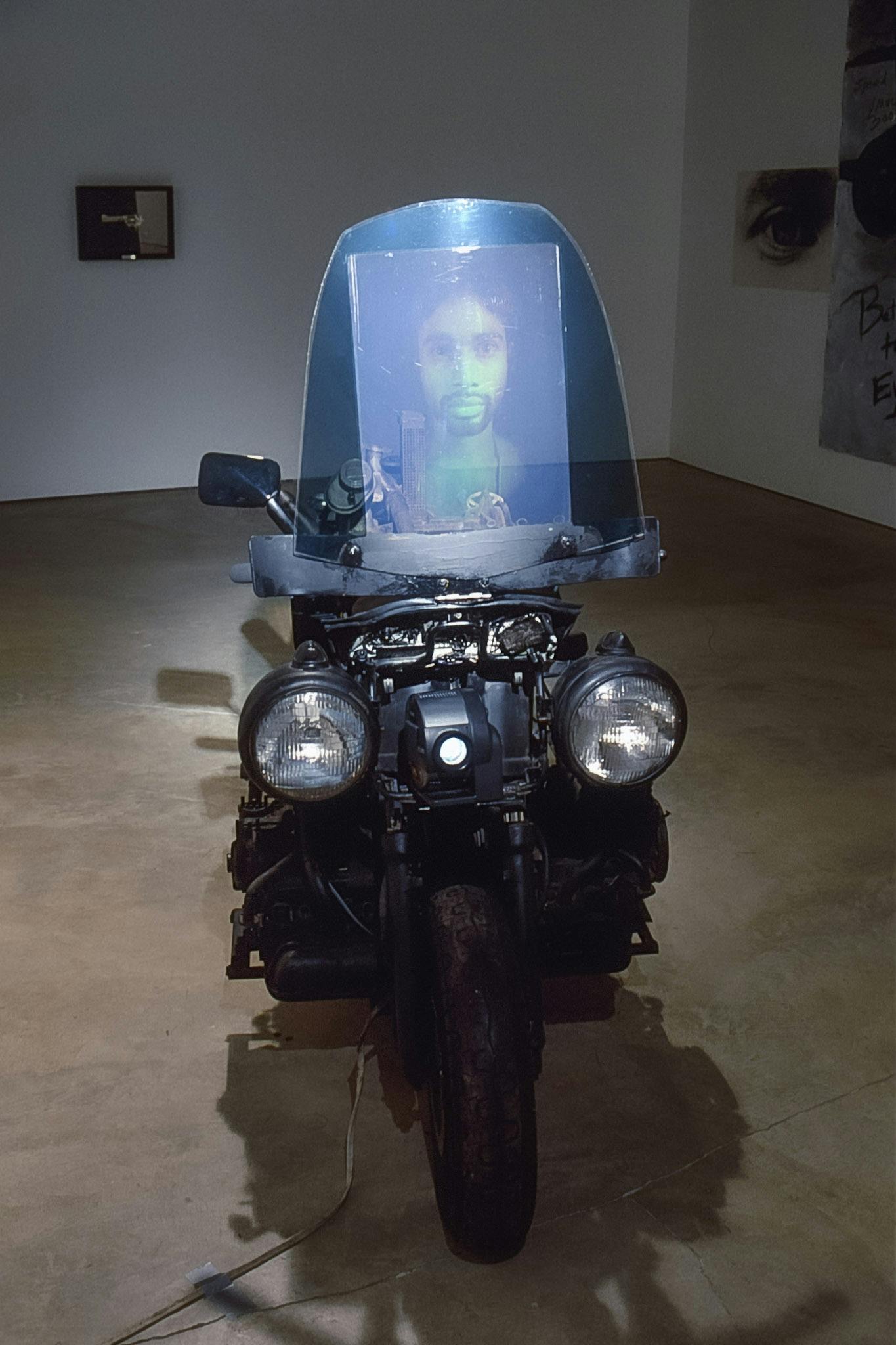 A black motorbike is installed in the gallery. An upside-down face shield is attached on top of the bike’s front, on which a man’s portrait photograph is attached. Handles are missing from this bike. 