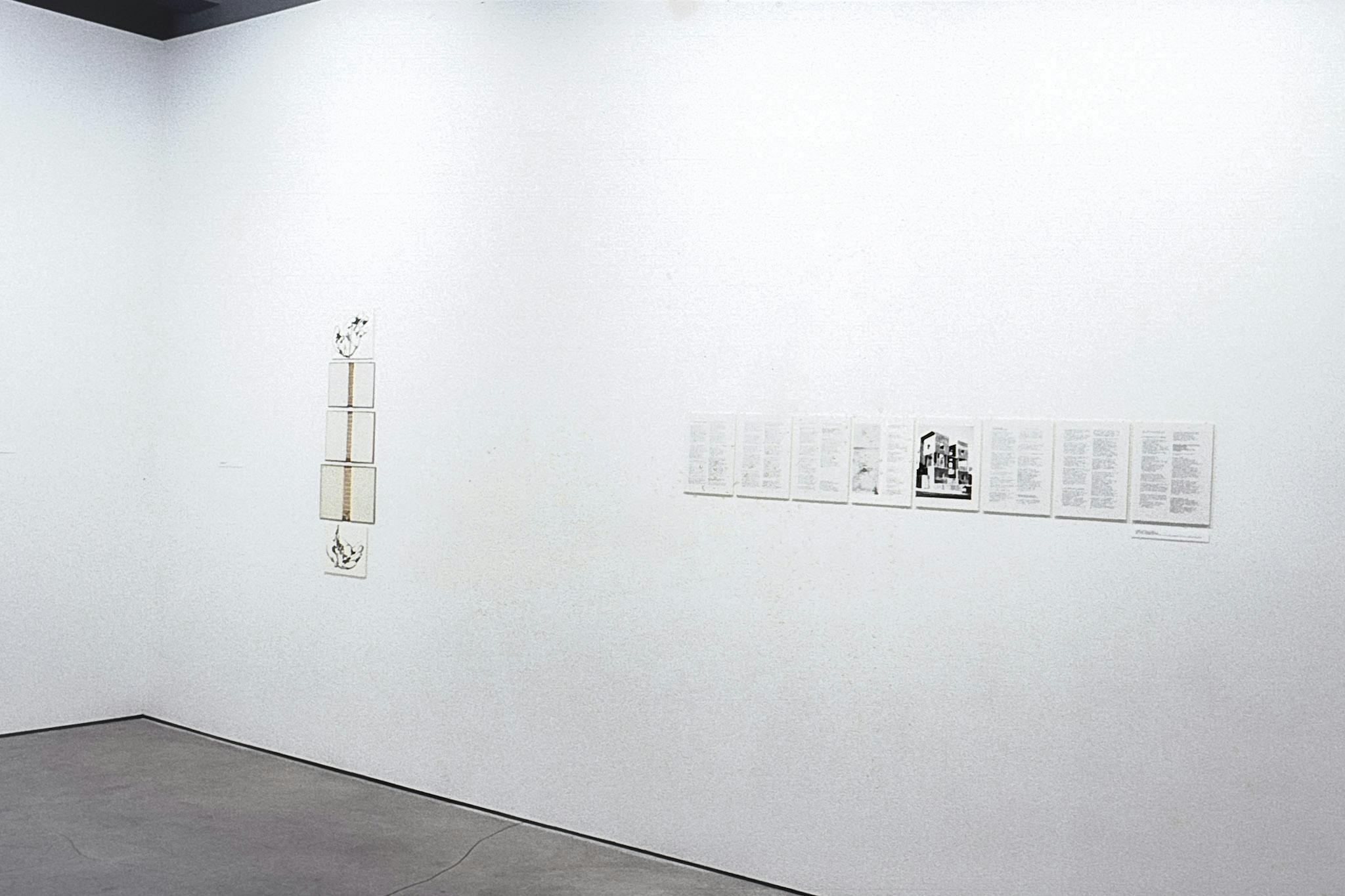 A closeup of a gallery wall with 2 artworks mounted on it. One shows 3 photos of a tall structure between two drawings of hands. The other is 7 panels of text with a photo of a building in the middle.