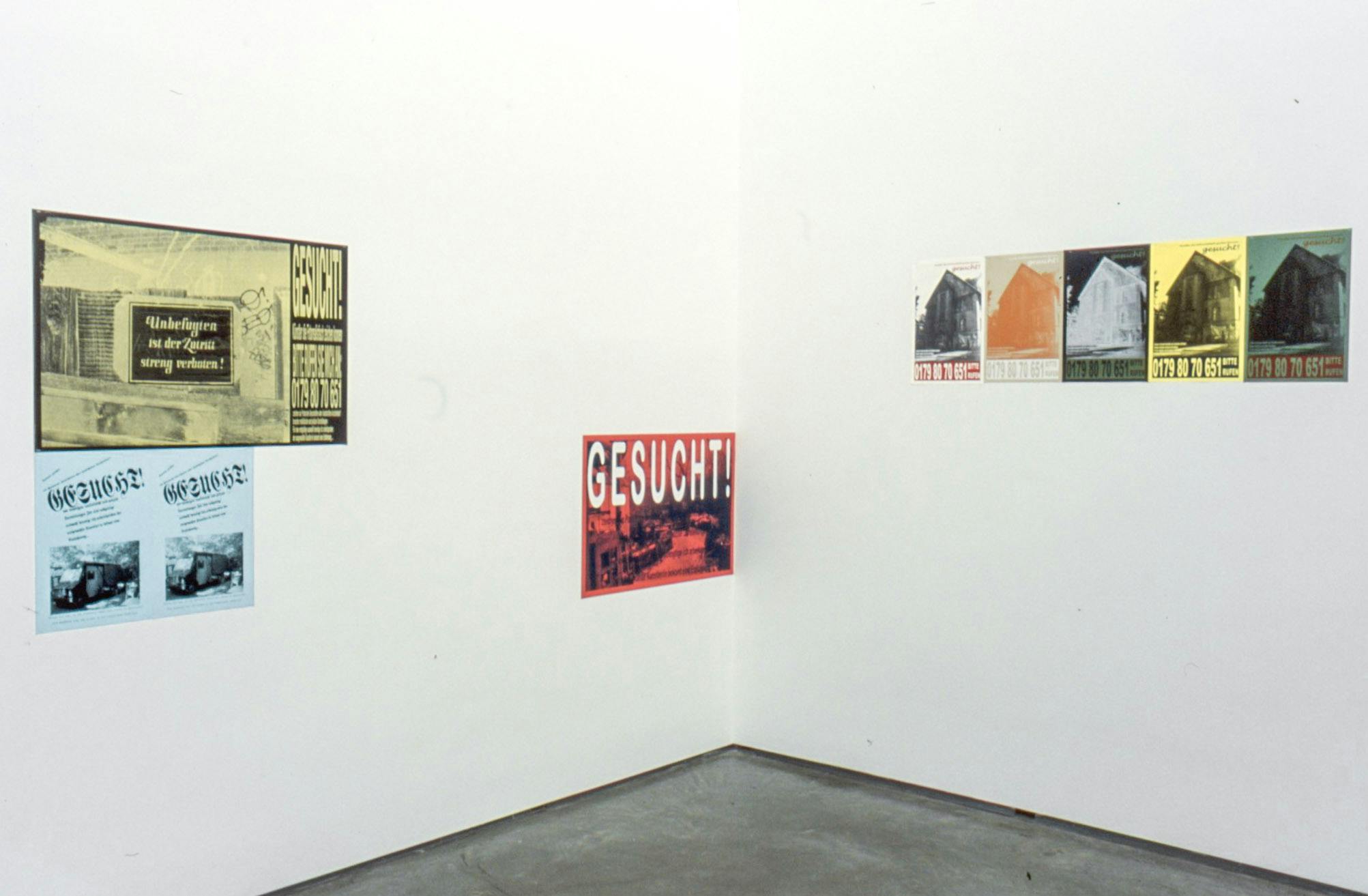 Installation image of posters. Five various-sized posters are mounted on the walls. The posters are coloured red, yellow, green, and light blue. Some of them are black and white. One poster shows the word “gesucht!” in all capital letters. 